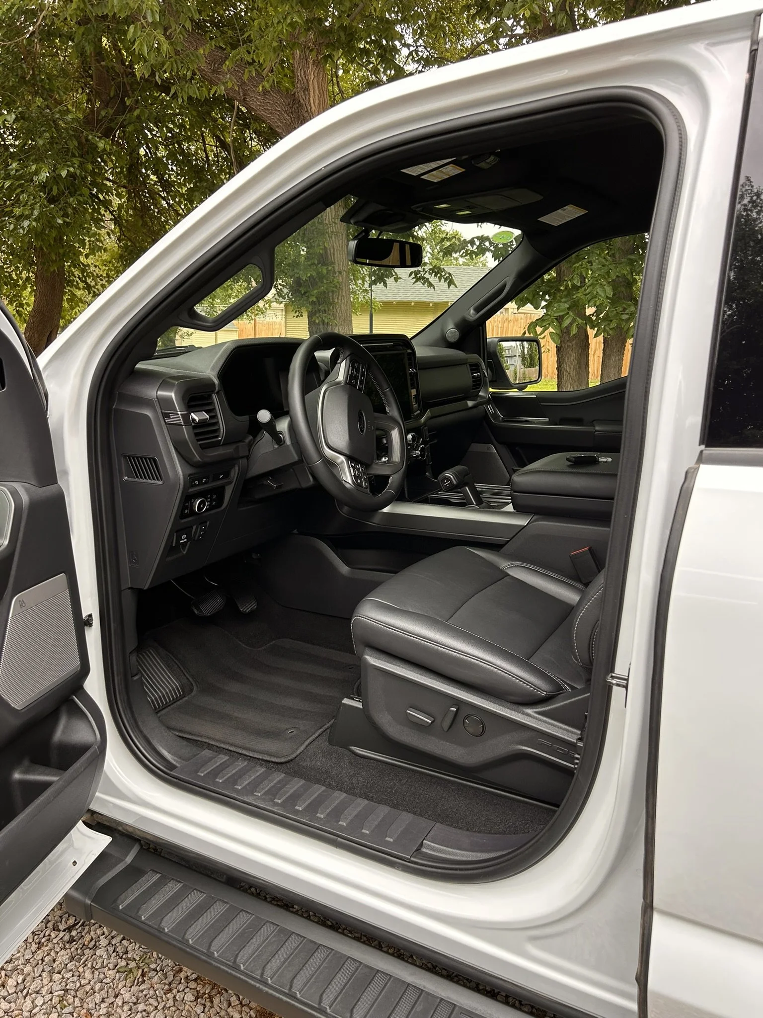 Interior view of a pickup truck's front passenger seat and dashboard, seen through the open door. The dashboard includes a steering wheel, center console, and various controls. The upholstery appears to be black, and there is a large side mirror reflecting a yard and trees outside.