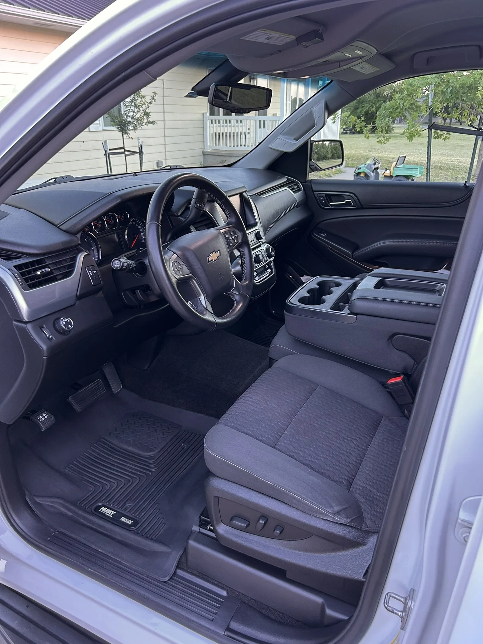 Interior of a Chevrolet pickup truck showing the dashboard with steering wheel, gear shift, center console with cup holders, and gray fabric seat, with a view through the passenger window of a small toy vehicle on the lawn outside.