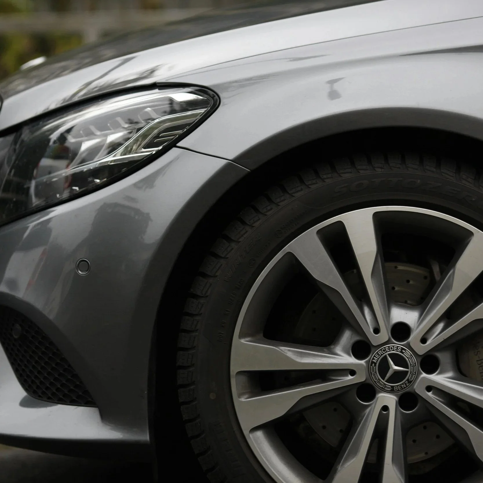 Close-up of the front left side of a silver Mercedes-Benz car, showing part of the headlight, front wheel, and tire.