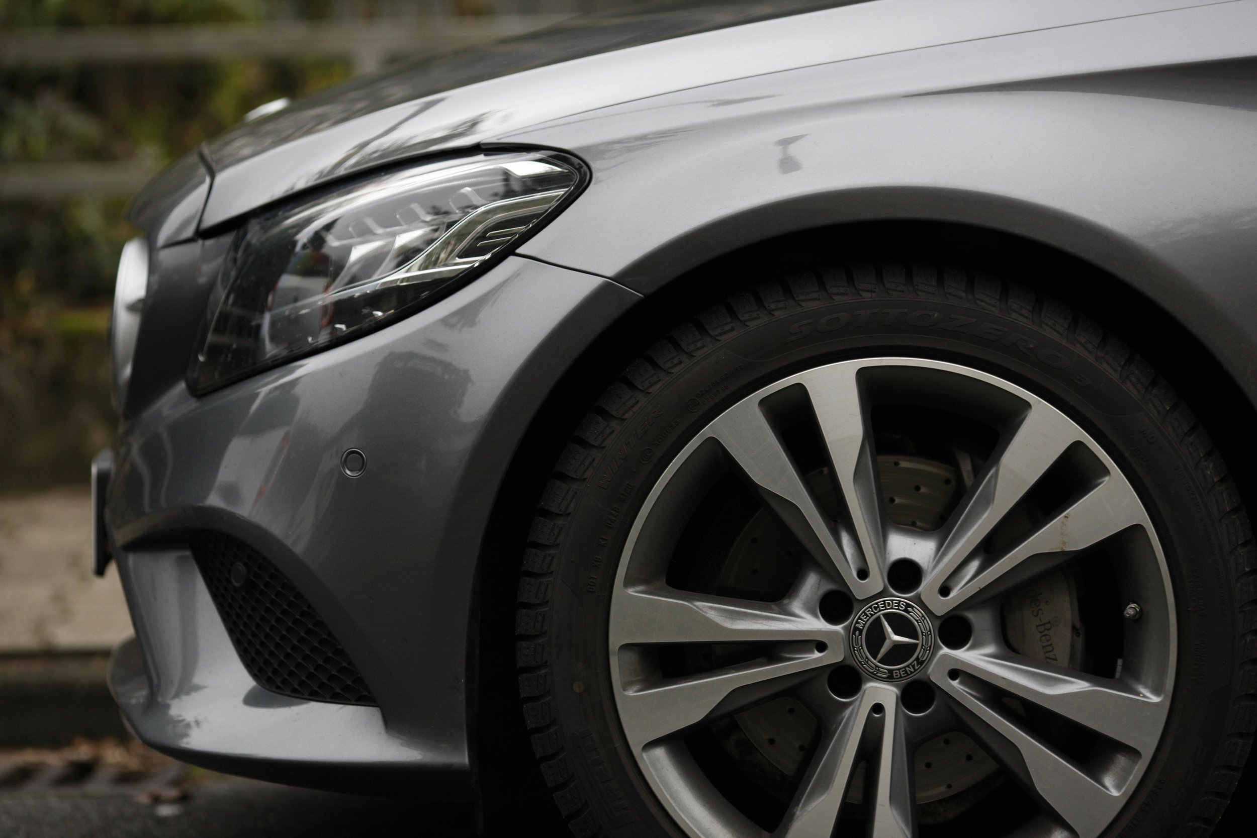 Close-up of the front left corner of a silver Mercedes-Benz car, showing the headlight, wheel, and part of the fender.