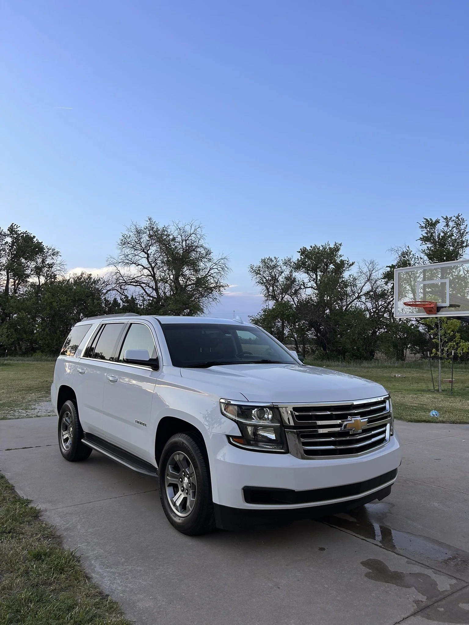 A white Chevrolet Tahoe SUV parked on a driveway next to a grassy area with trees under a clear blue sky with some clouds.
