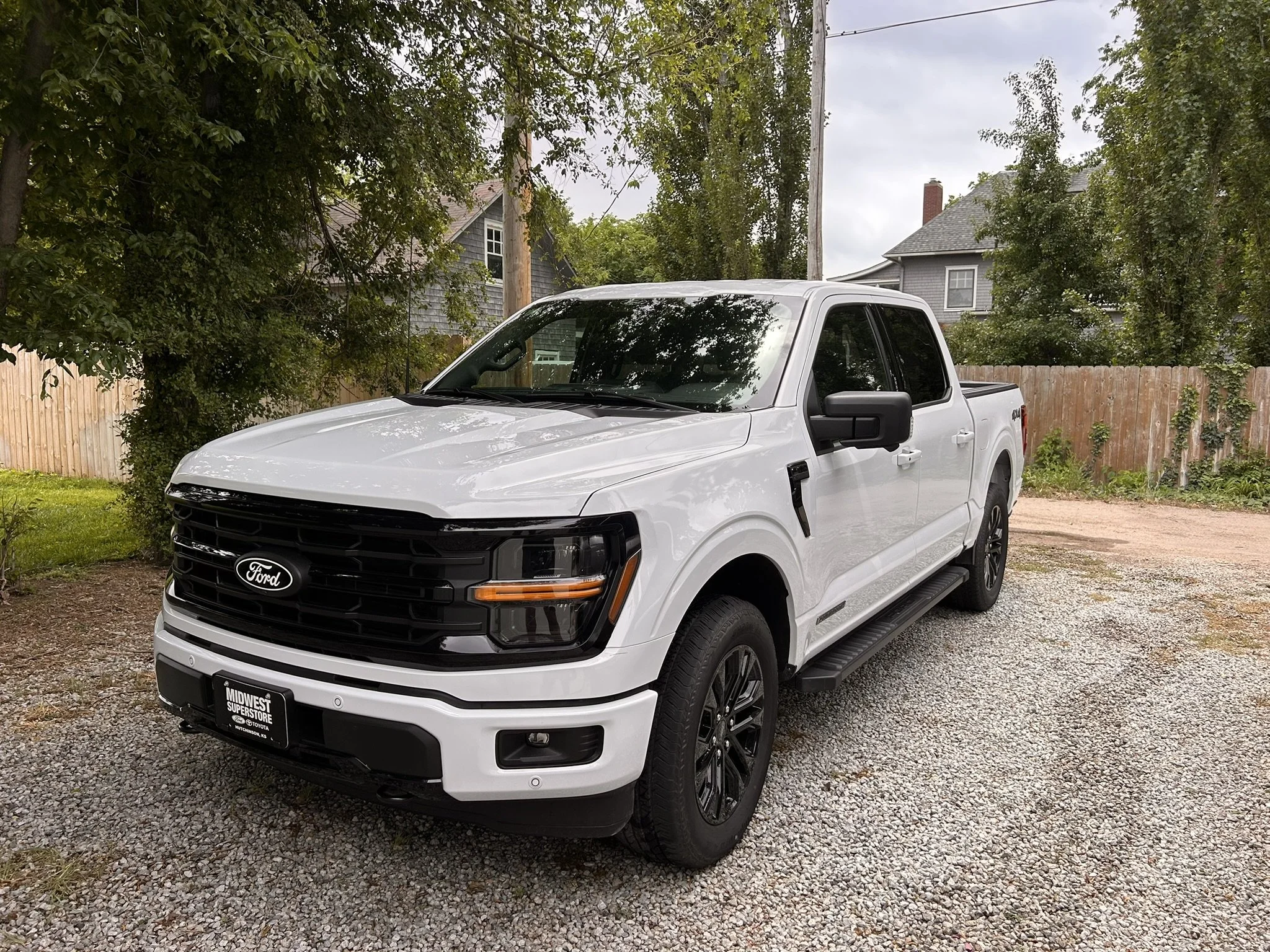 A white Ford pickup truck parked on a gravel driveway, with a wooden fence and trees in the background. There is a gray house with a sloped roof visible behind the truck.