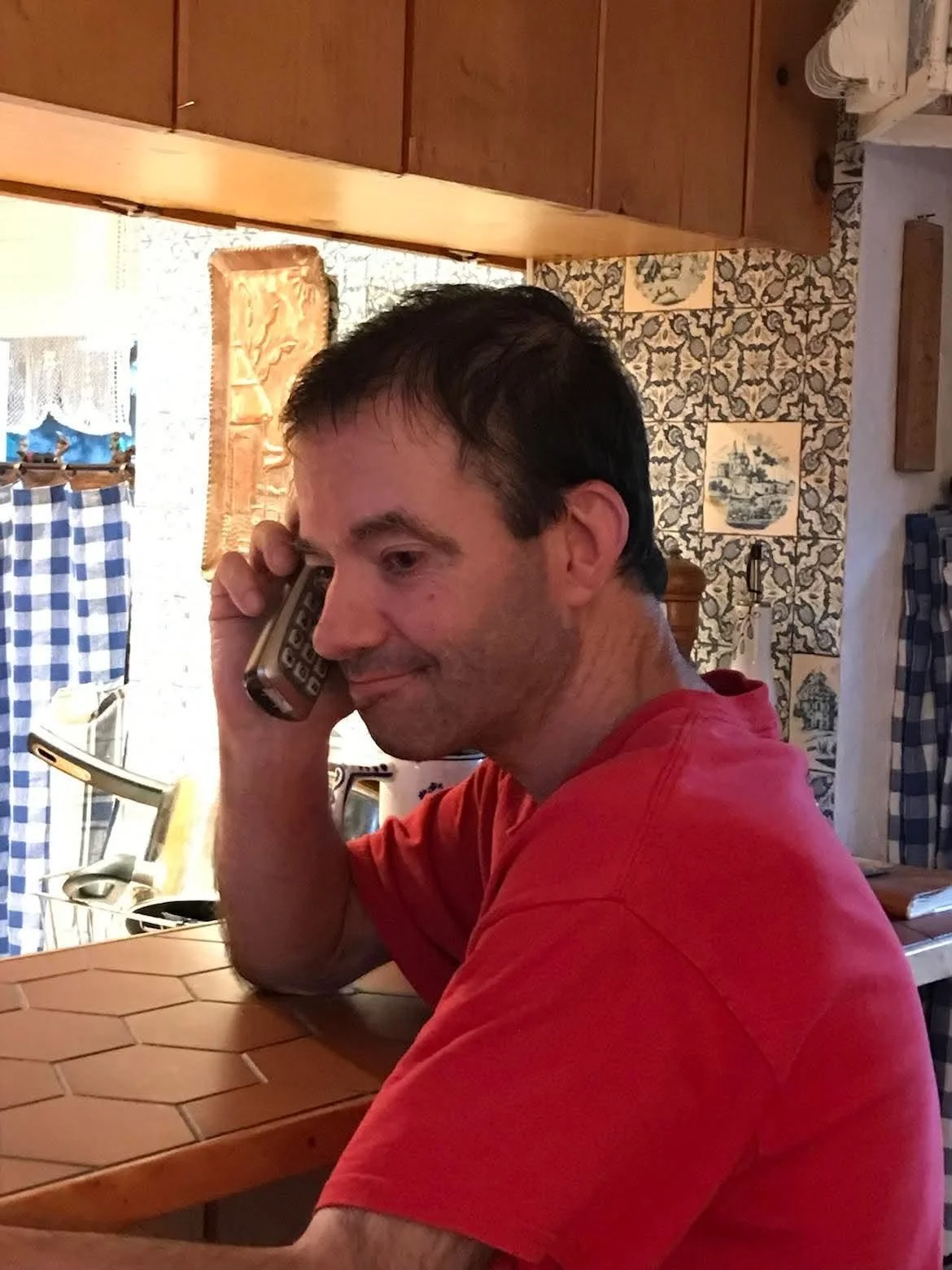Un homme parle au téléphone dans une cuisine aux murs décorés de carreaux, portant un t-shirt rouge.