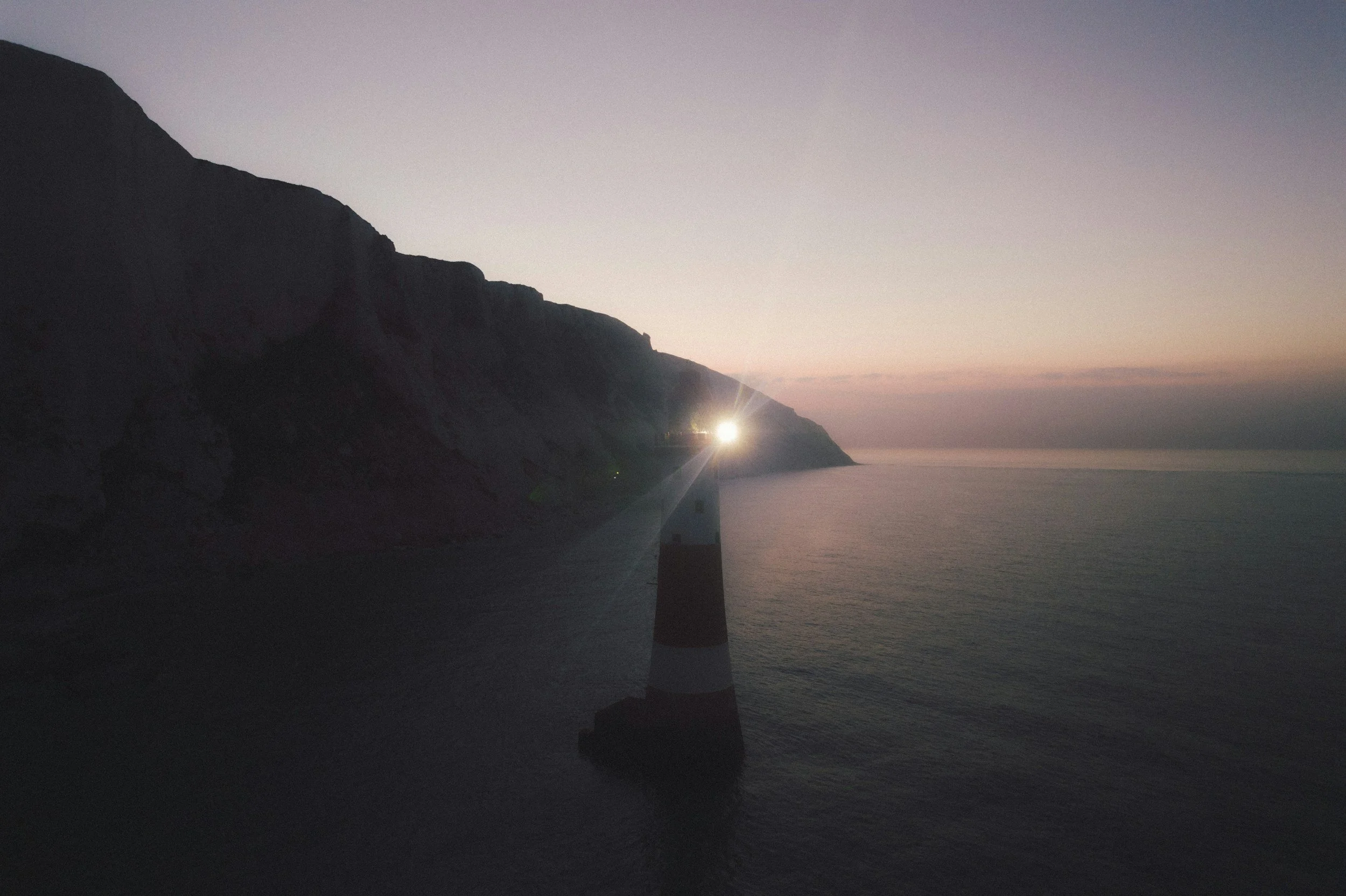 A lighthouse on a rocky coastline at dusk with a faint glow on the horizon, calm ocean waters, and mountain cliffs on the left.