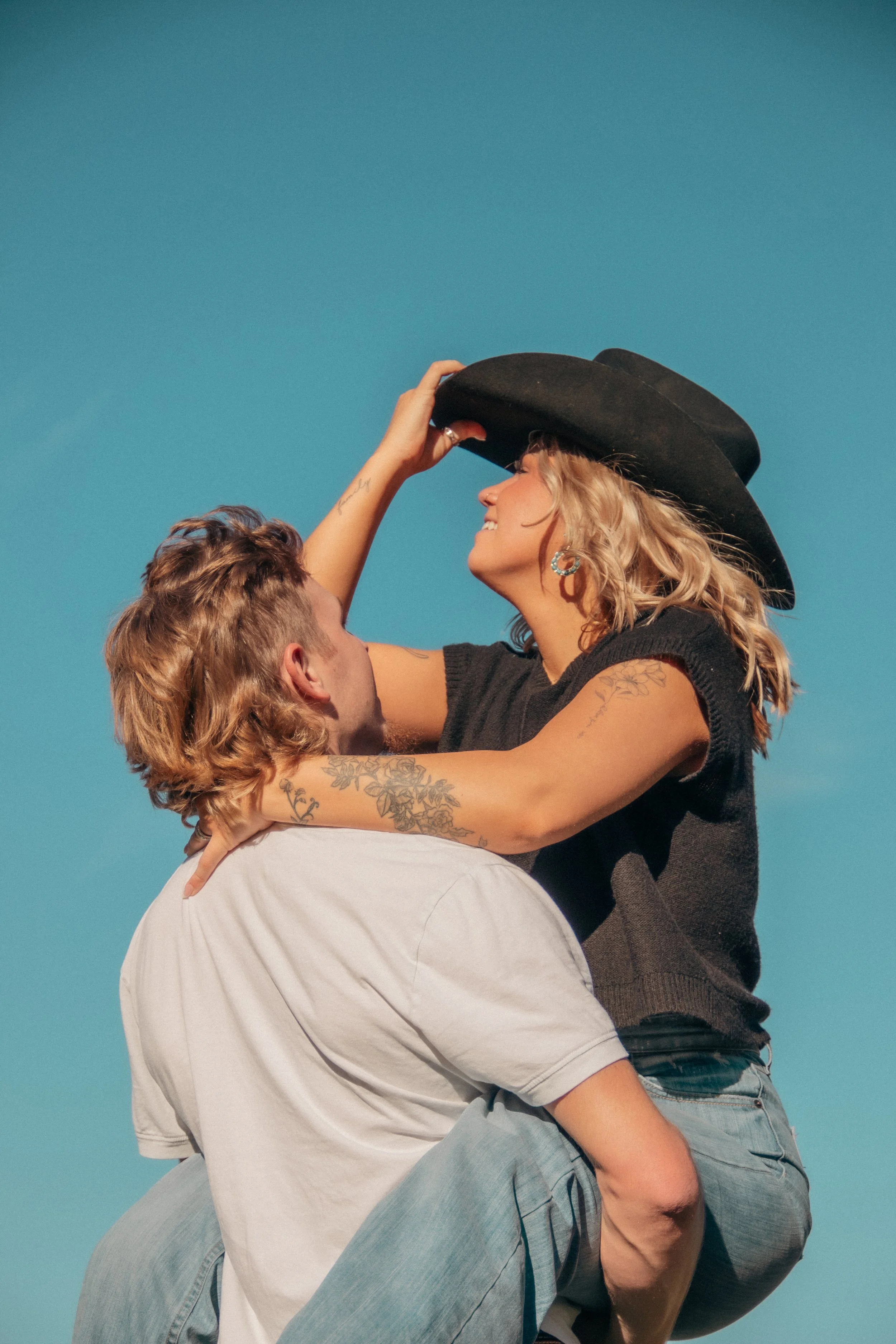A woman wearing a black hat and earrings, smiling, being lifted by a man, against a bright blue sky.