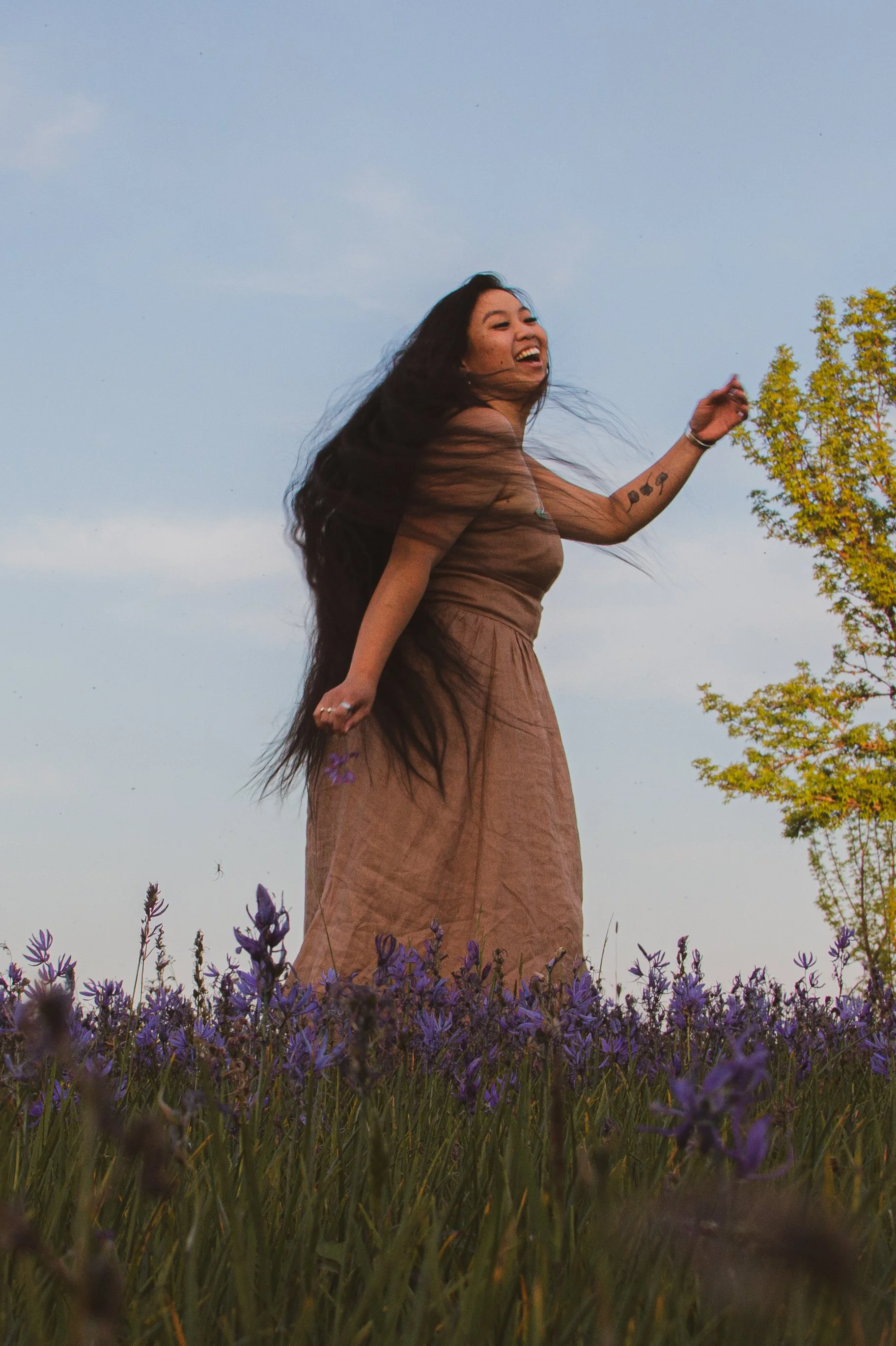 A woman with long dark hair and multiple tattoos on her arm standing in a lavender field, smiling and looking to her right against a blue sky background.