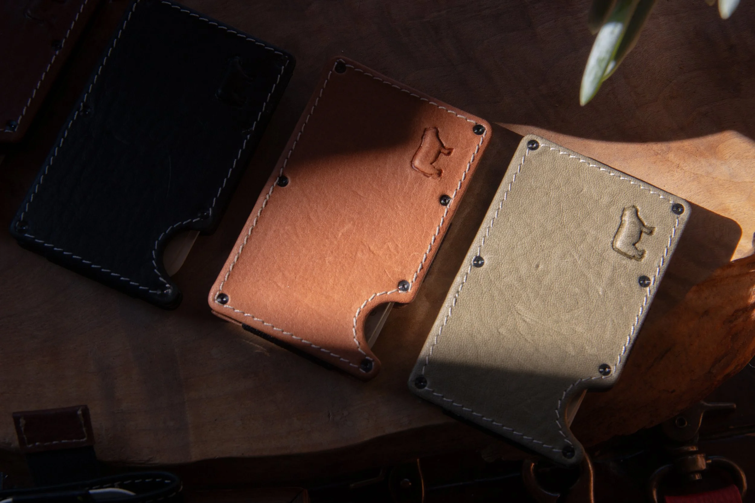 Close-up of three leather cardholders in black, brown, and beige placed on a wooden surface with a plant in the background.