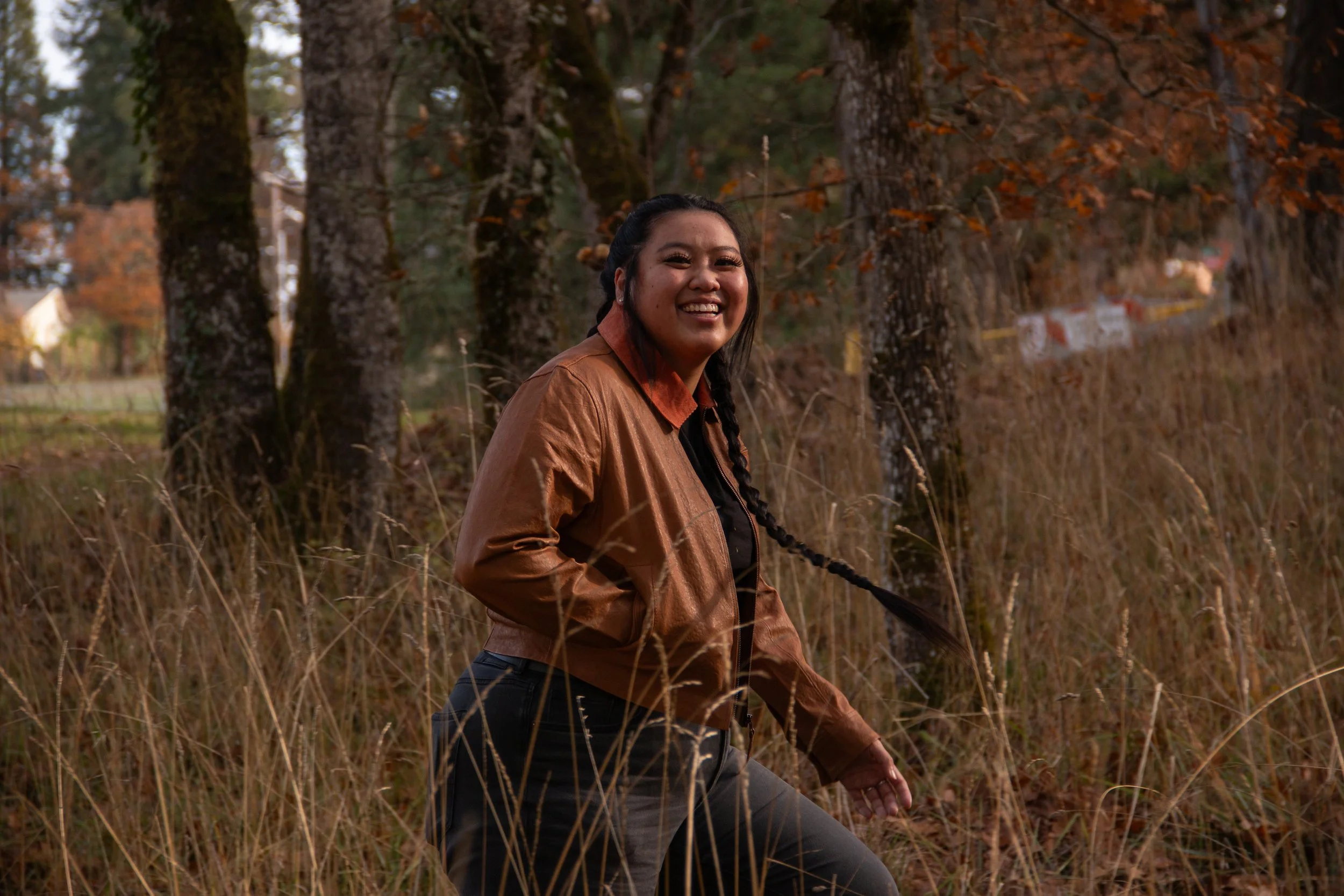 A smiling woman with long dark hair tied in a braid, wearing a brown jacket and black pants, standing outdoors in a grassy area with trees and leaves in fall colors.