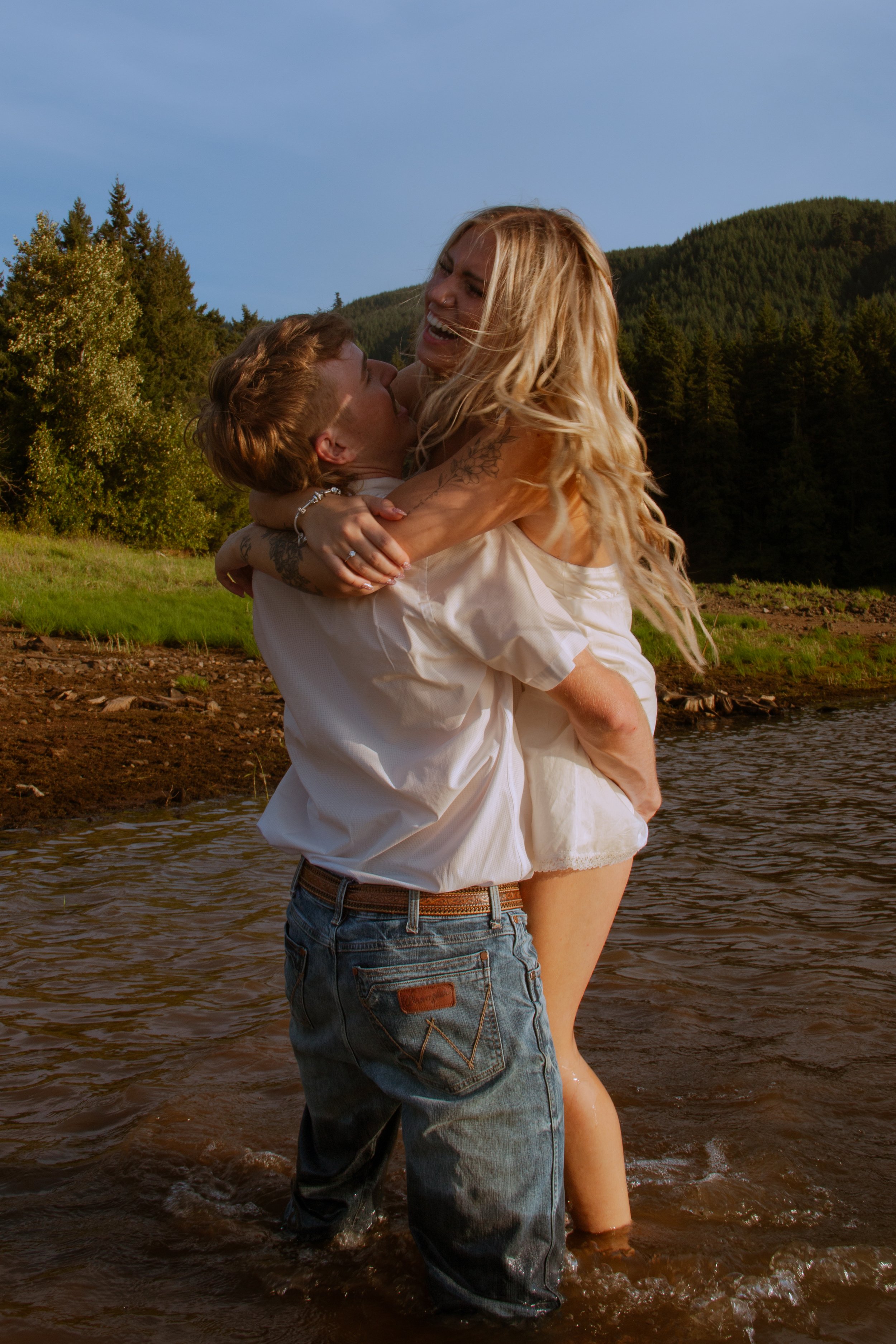 A man is holding a woman in a river, both smiling, with trees and mountains in the background on a clear day.
