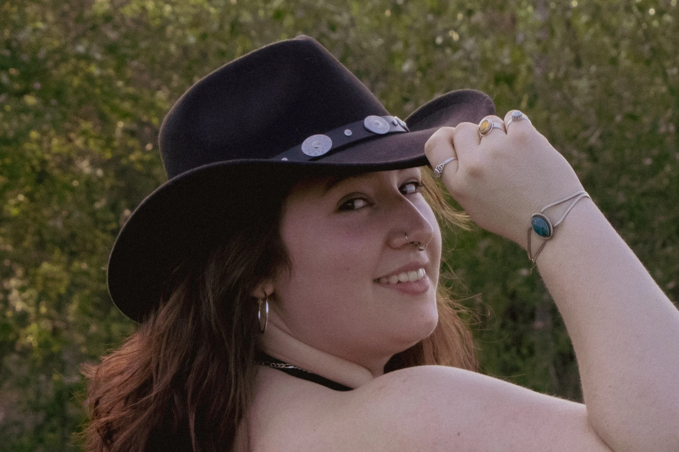 A woman with long reddish hair and piercings, smiling, wearing a brown cowboy hat decorated with coins, outdoors with green trees in the background.