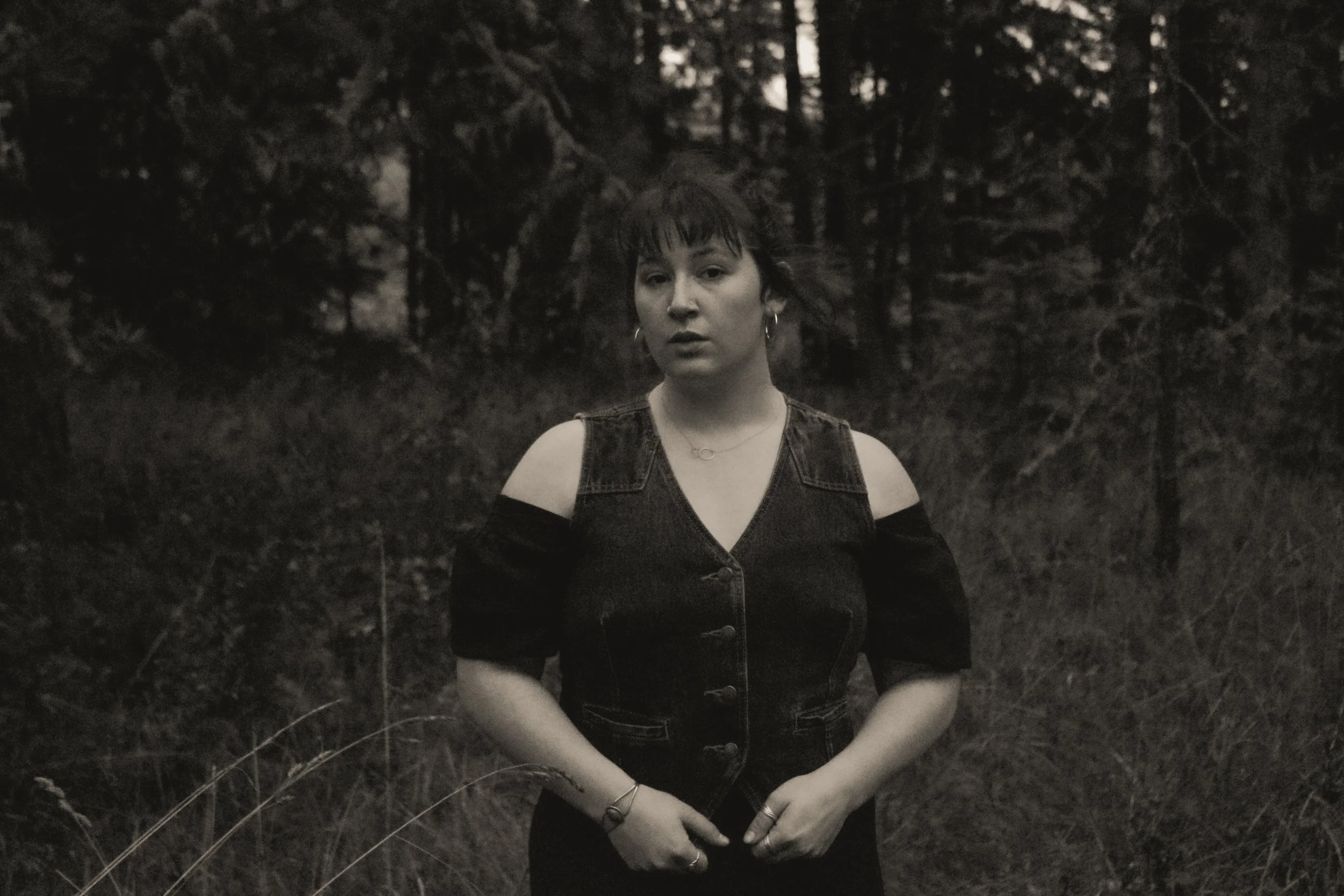 A person stands outdoors in a forested area during dusk or night. They have short, dark hair with bangs, and are wearing a sleeveless denim vest with black straps, earrings, and jewelry. Their expression is neutral, and they are looking directly at the camera.