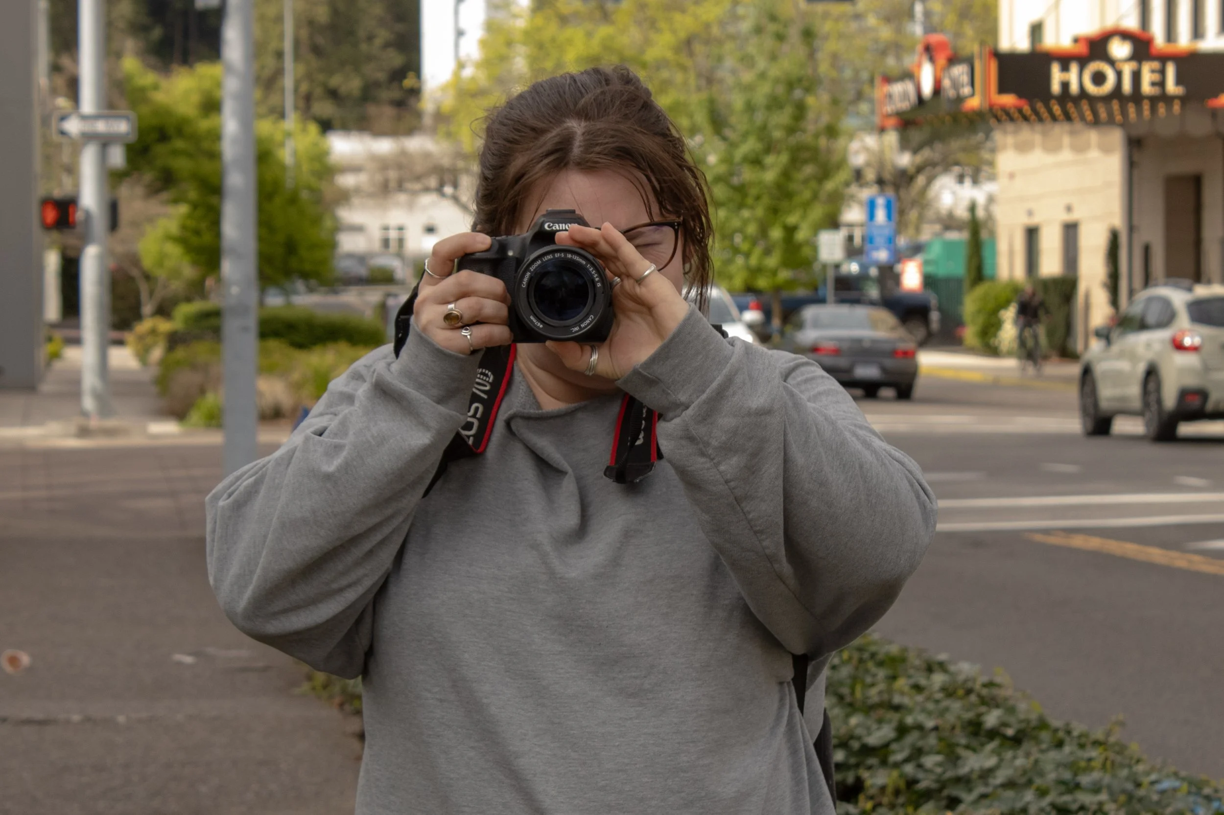 A person with shoulder-length brown hair and glasses, wearing a gray hoodie, holding a Canon camera up to their face and taking a photo outdoors in an urban setting with cars, trees, and a building in the background.
