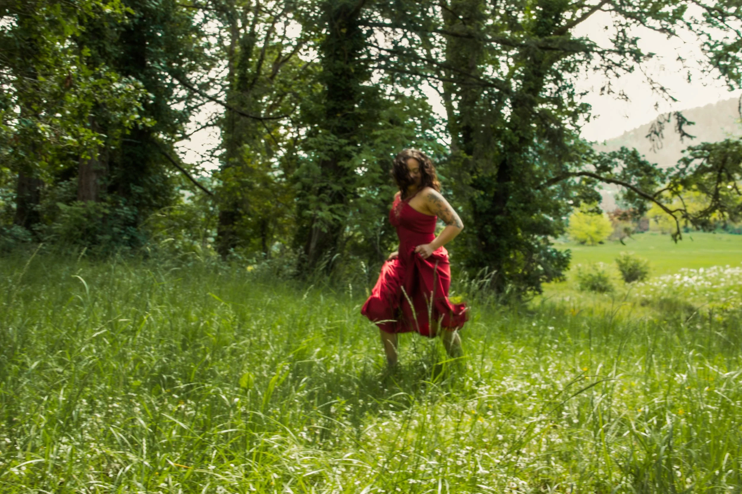 A woman in a red dress standing in a green grassy field surrounded by trees, with her hands on her hips and looking downward.
