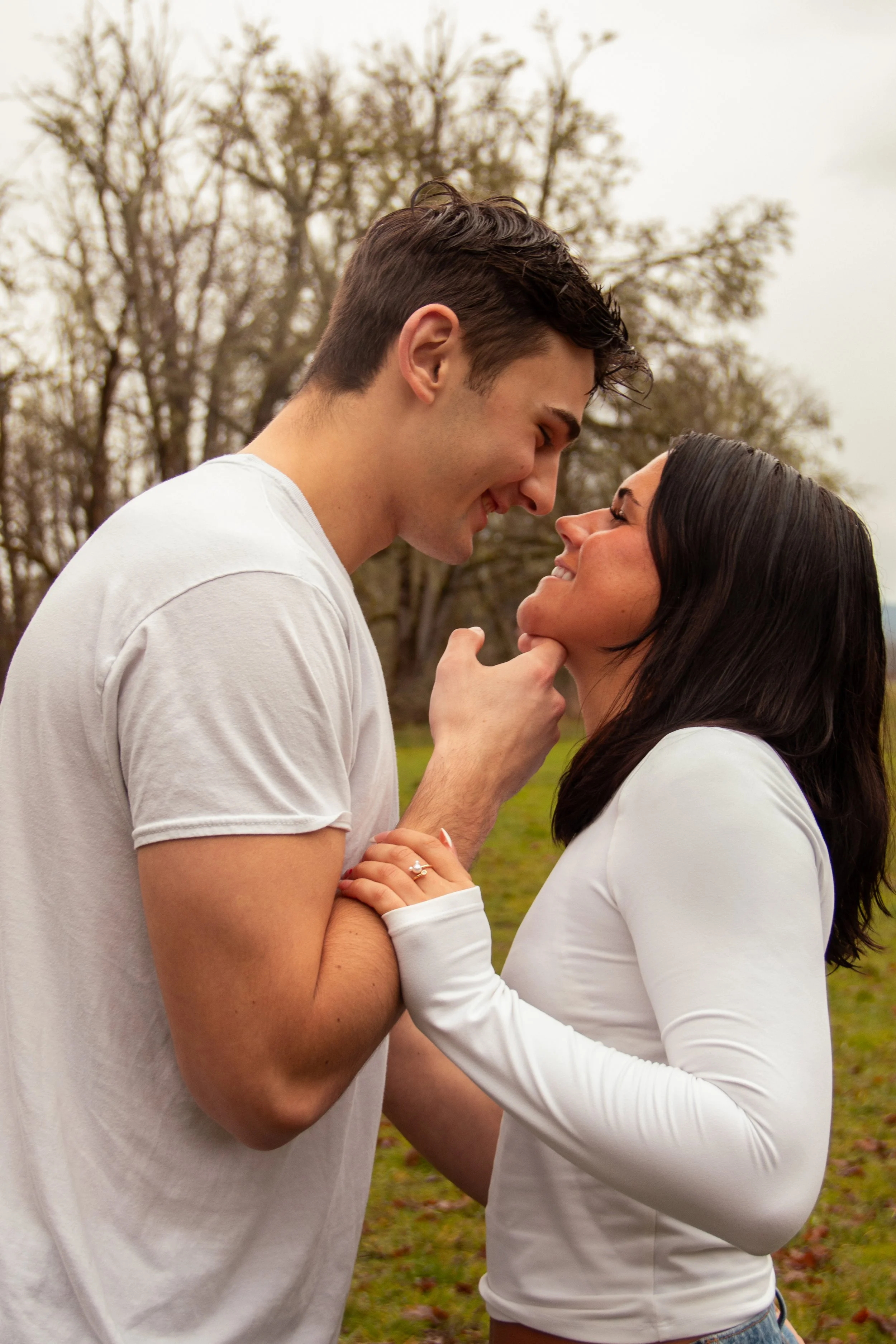 A young man and woman are close together outdoors, smiling and looking into each other's eyes, with their foreheads touching and hands clasped.