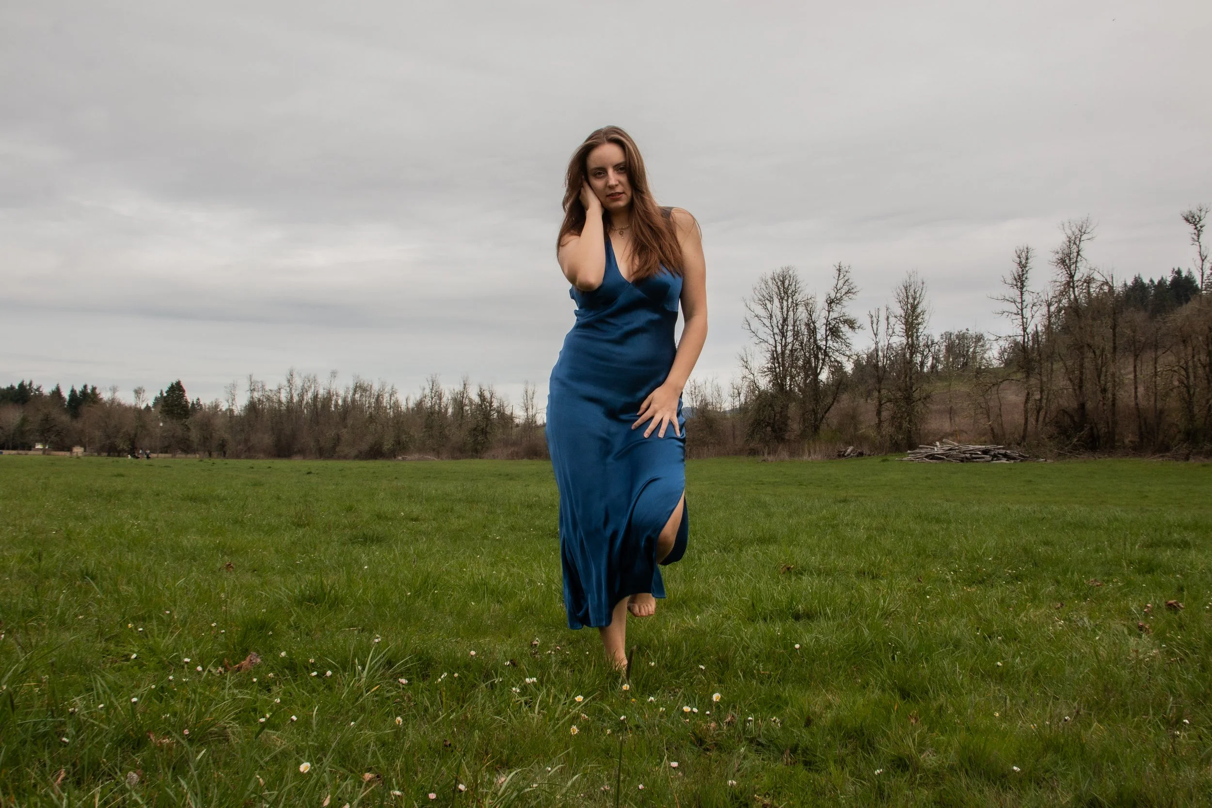A young woman with long hair wearing a blue dress walking barefoot across a grassy field on a cloudy day.