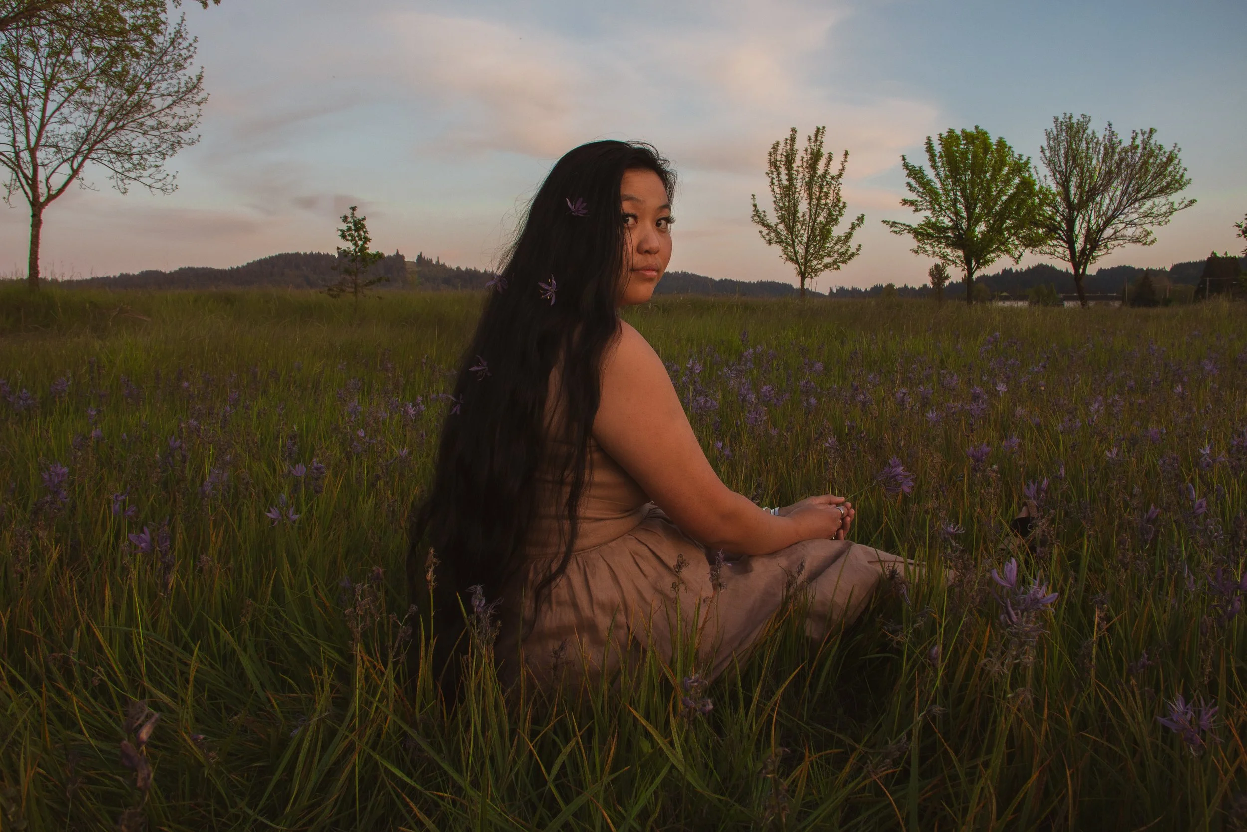 A young woman with long dark hair and purple flowers in her hair, sitting in a field of purple flowers, with trees and hills in the background during sunset.