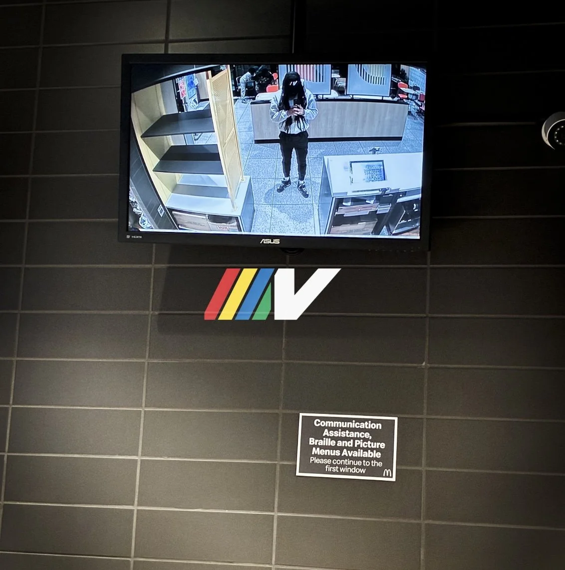 A security monitor showing a person taking a photo with a phone in a store or fast-food restaurant. The person is wearing a face mask, jacket, and pants. The monitor is mounted on a tiled wall, with a McDonald's sign below it and a sign providing communication assistance and menu options.