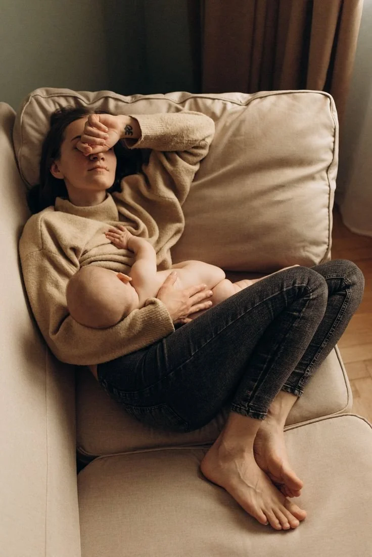 A woman with dark hair lying on a beige sofa with a baby resting on her chest, holding the baby with one hand while covering her eyes with her other arm, in a cozy living room.