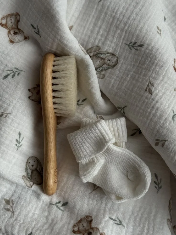 A soft baby blanket with teddy bear and bunny patterns, a small wooden-handled brush, and a pair of white baby socks laid on top.