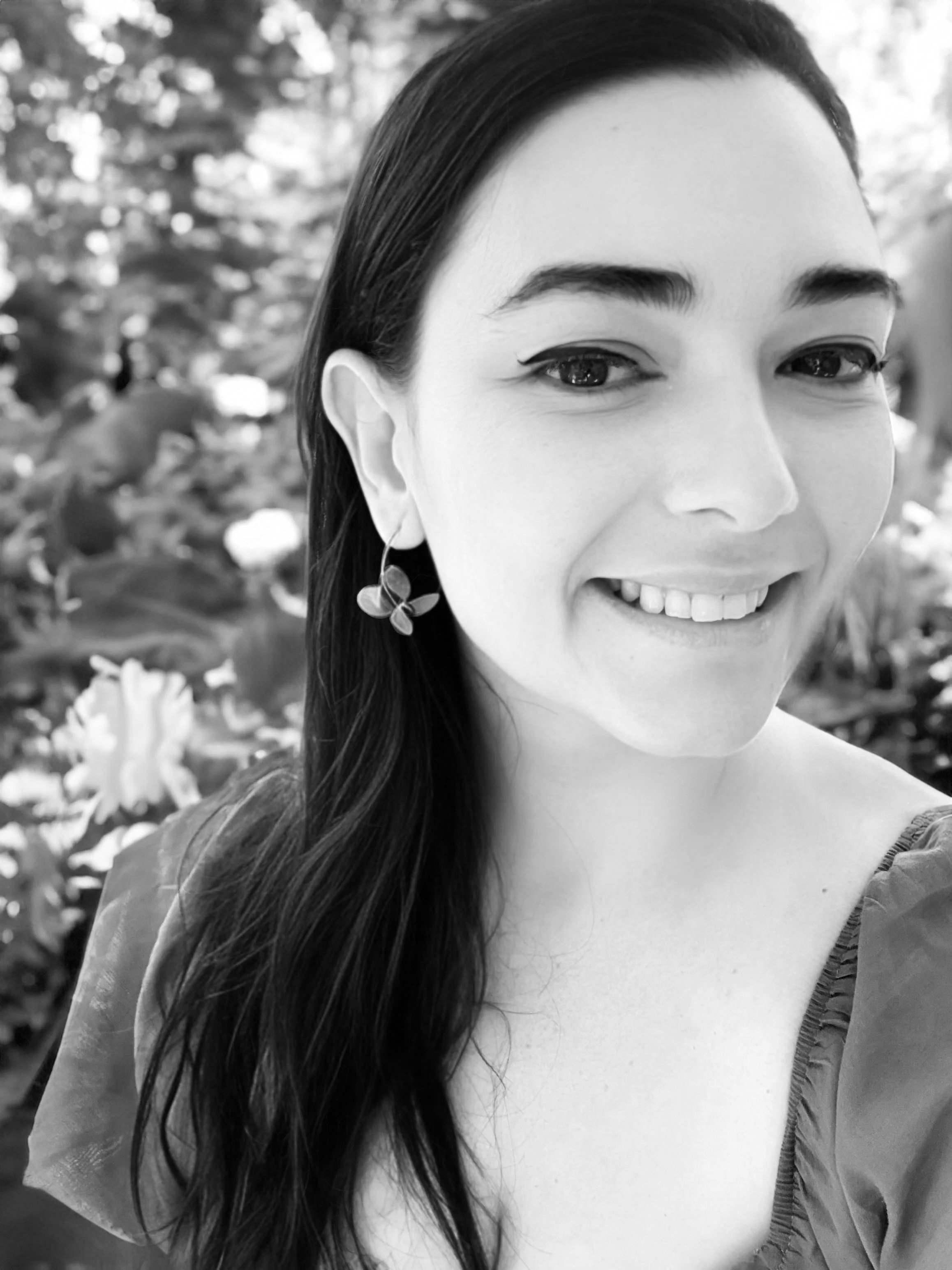 Close-up of a smiling woman with long dark hair, wearing earrings, in front of a blurred floral background.