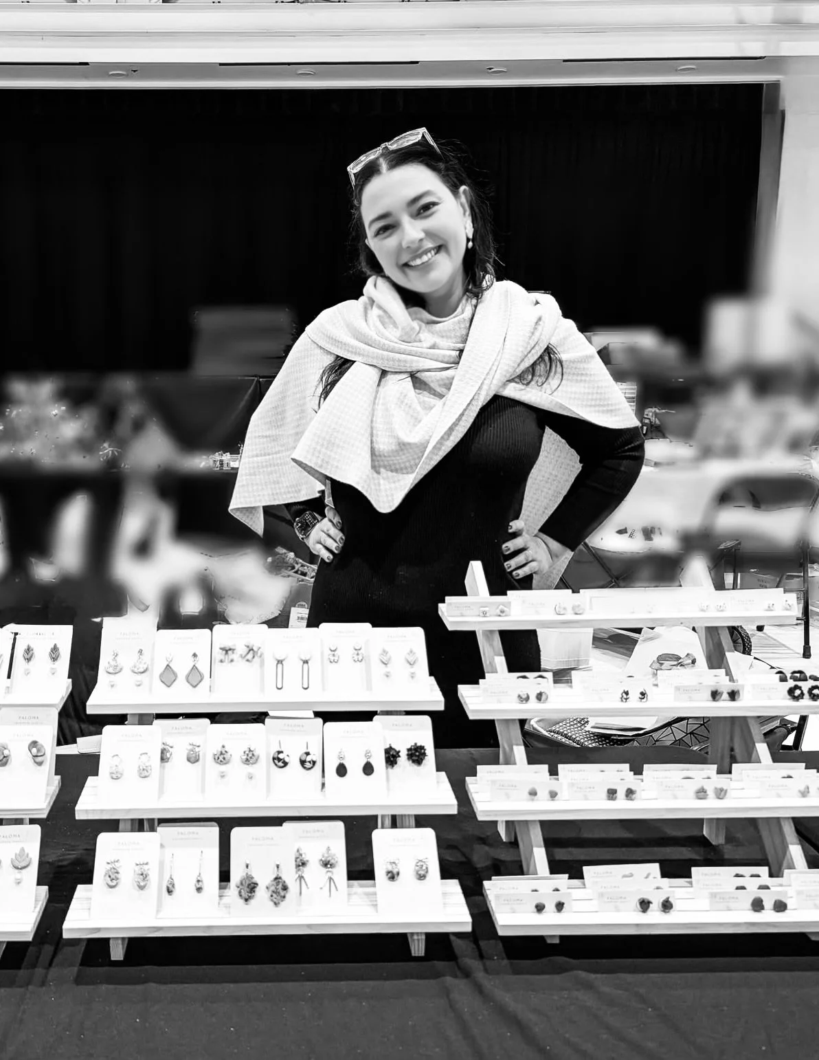 A woman with glasses on her head and earrings stands behind a jewelry display table, smiling at the camera. The table has various earrings and jewelry pieces arranged on display stands.