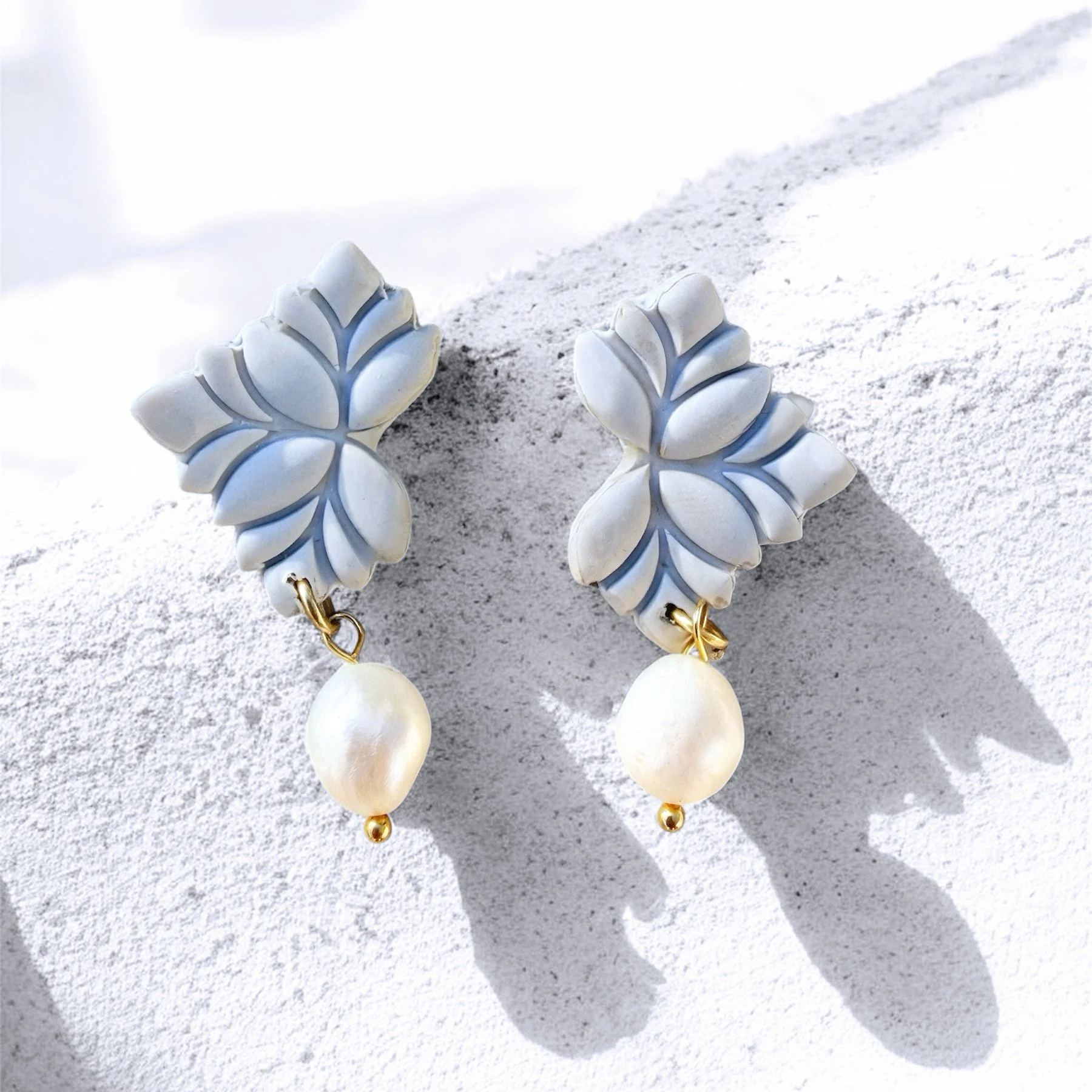 A pair of white earrings with leaf-shaped top pieces and large oval pearls hanging from small gold beads, placed on a textured white surface with shadows.