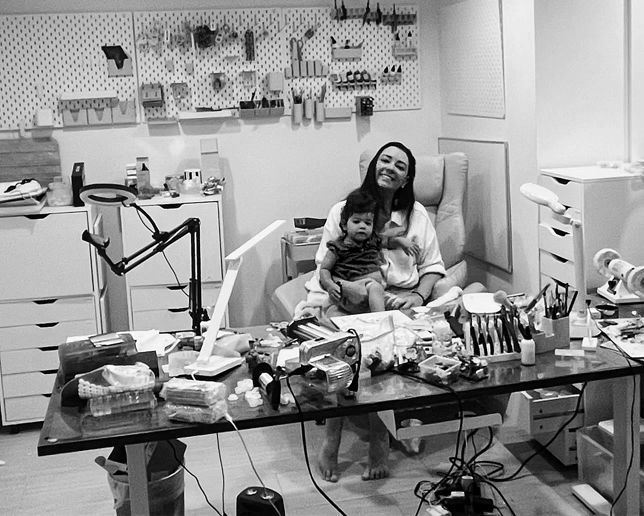 A woman smiling and sitting in a dental office with a young child on her lap, surrounded by dental tools, supplies, and equipment on a cluttered table.
