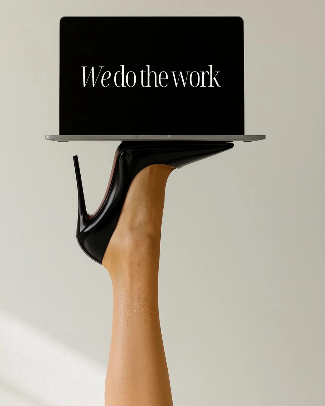 A hand wearing a black high heel shoe is holding a laptop with the message "We do the work" on the screen.