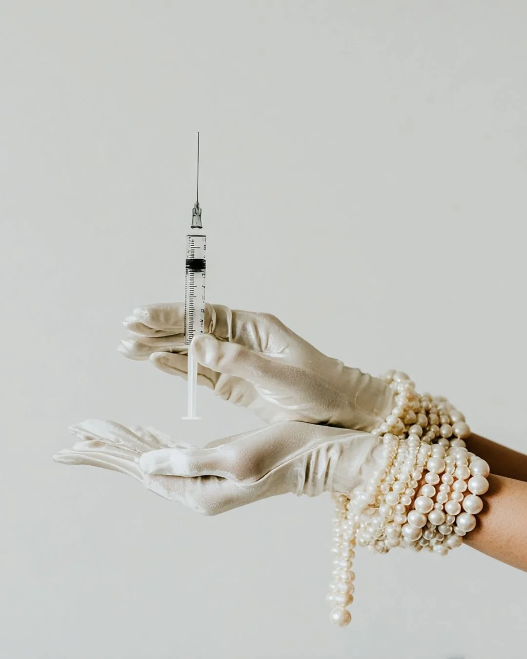 A hand wearing pearl jewelry holding a syringe with a needle against a plain background.