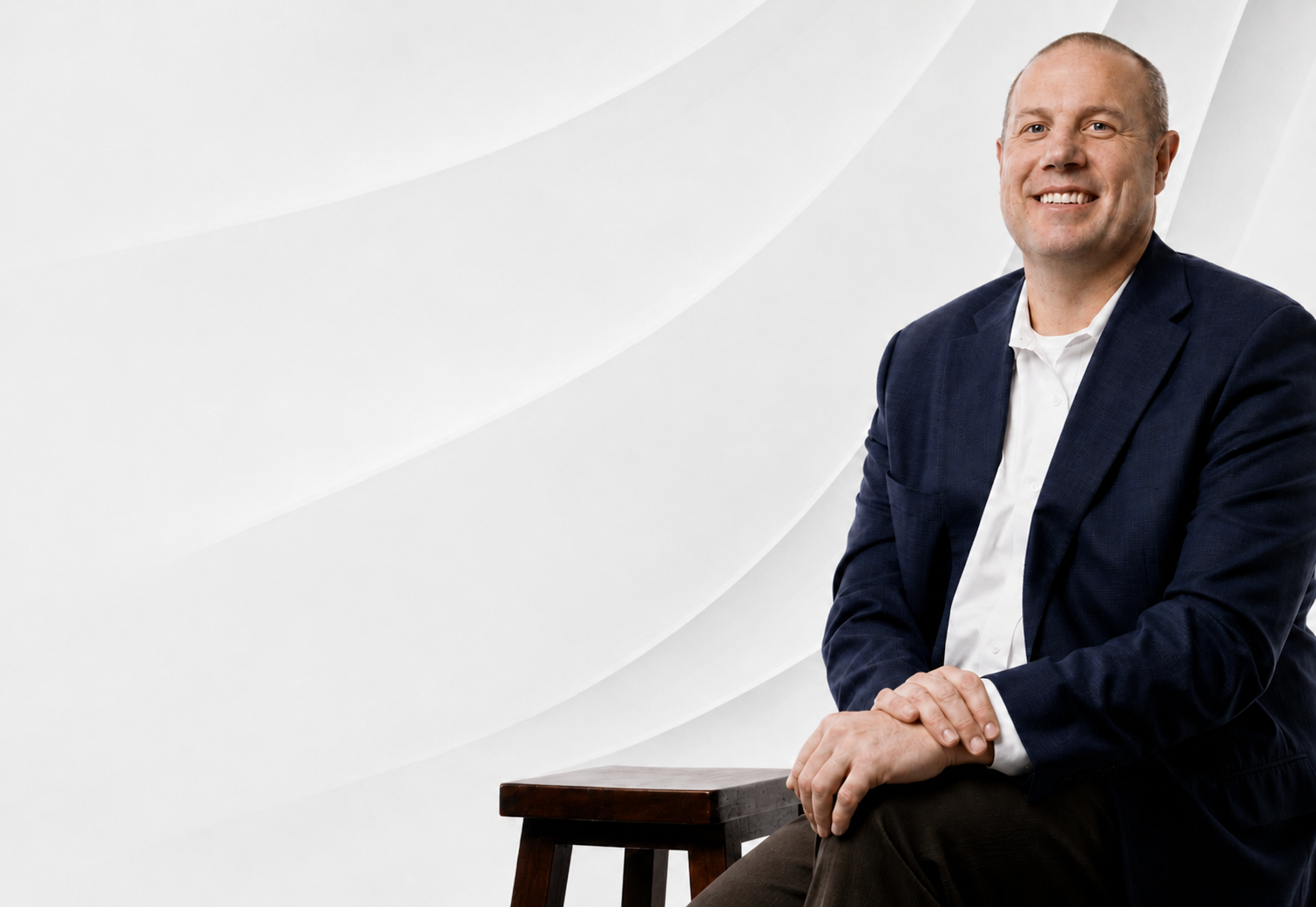 A man in a dark blue blazer and white shirt sitting on a stool against a white background with subtle curved lines.