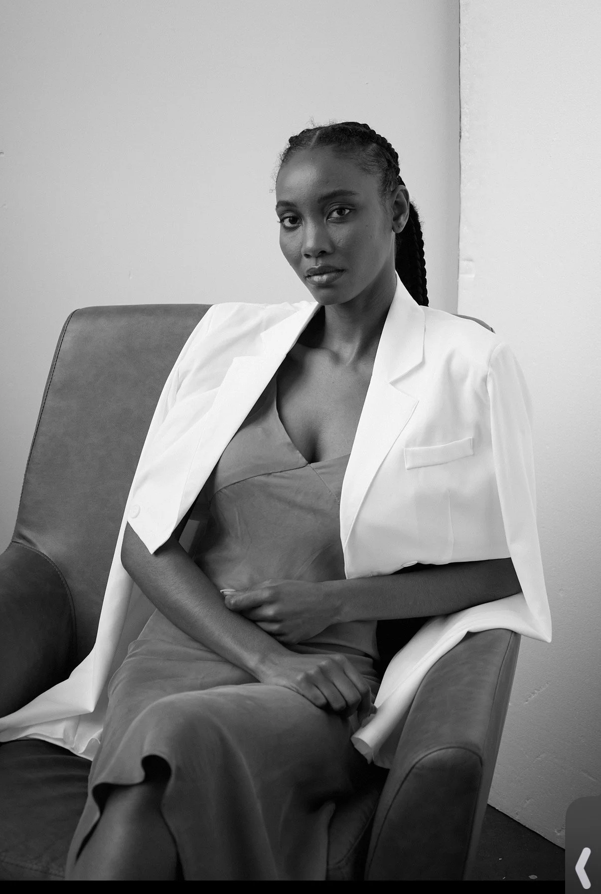A woman with braided hair, wearing a light-colored blazer over a dress, sitting on an armchair against a plain wall.