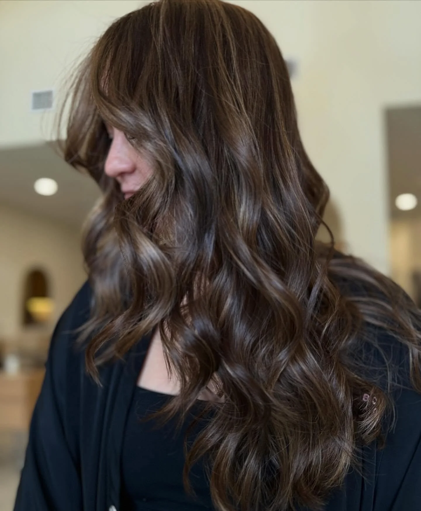 Woman with long, wavy brown hair, styled with loose curls, wearing a black top in an indoor setting.