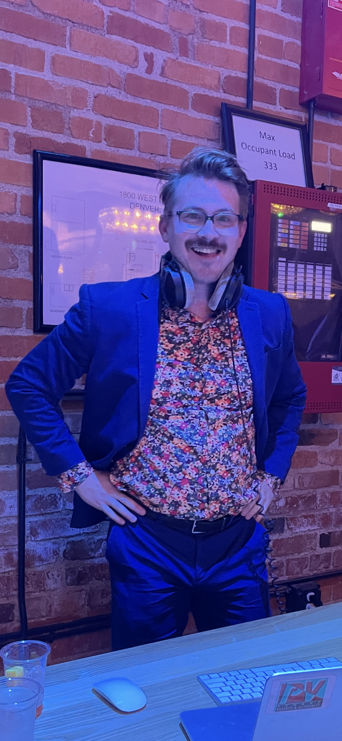 A man wearing glasses, a floral shirt, and a blue blazer standing with hands on hips, smiling, in front of a brick wall with framed maps and a fire alarm panel.