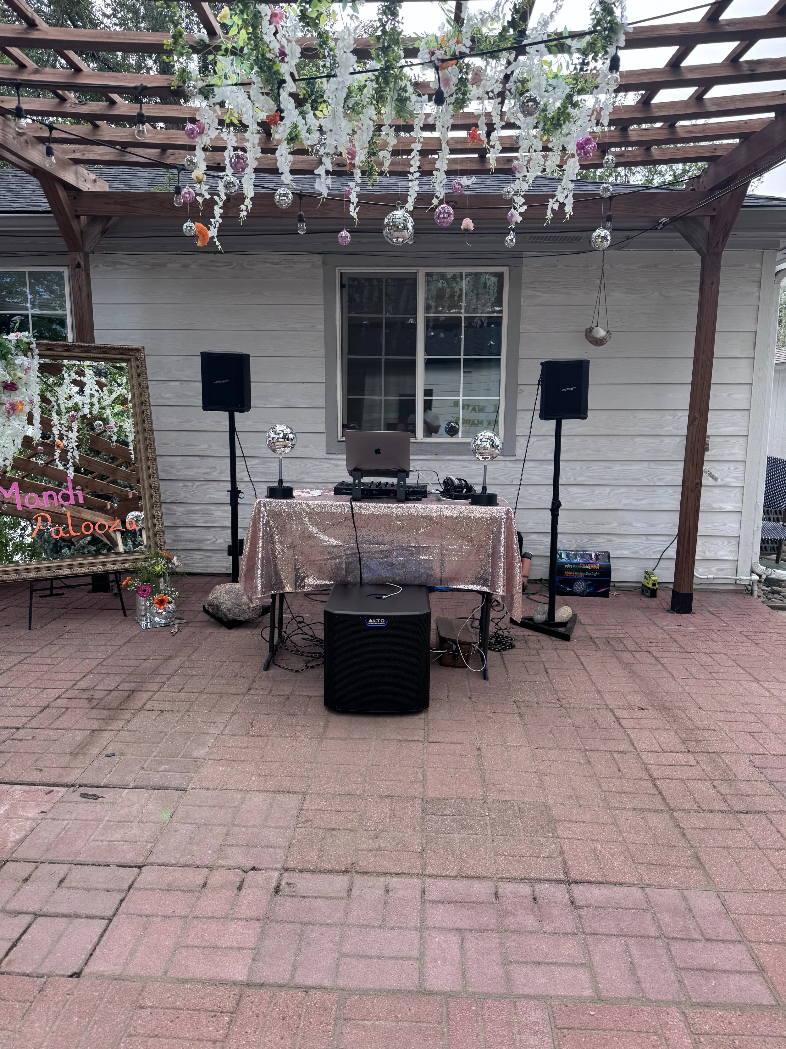 Outdoor DJ setup with table, speakers, subwoofer, laptop, and DJ controller with decorations around the table.
