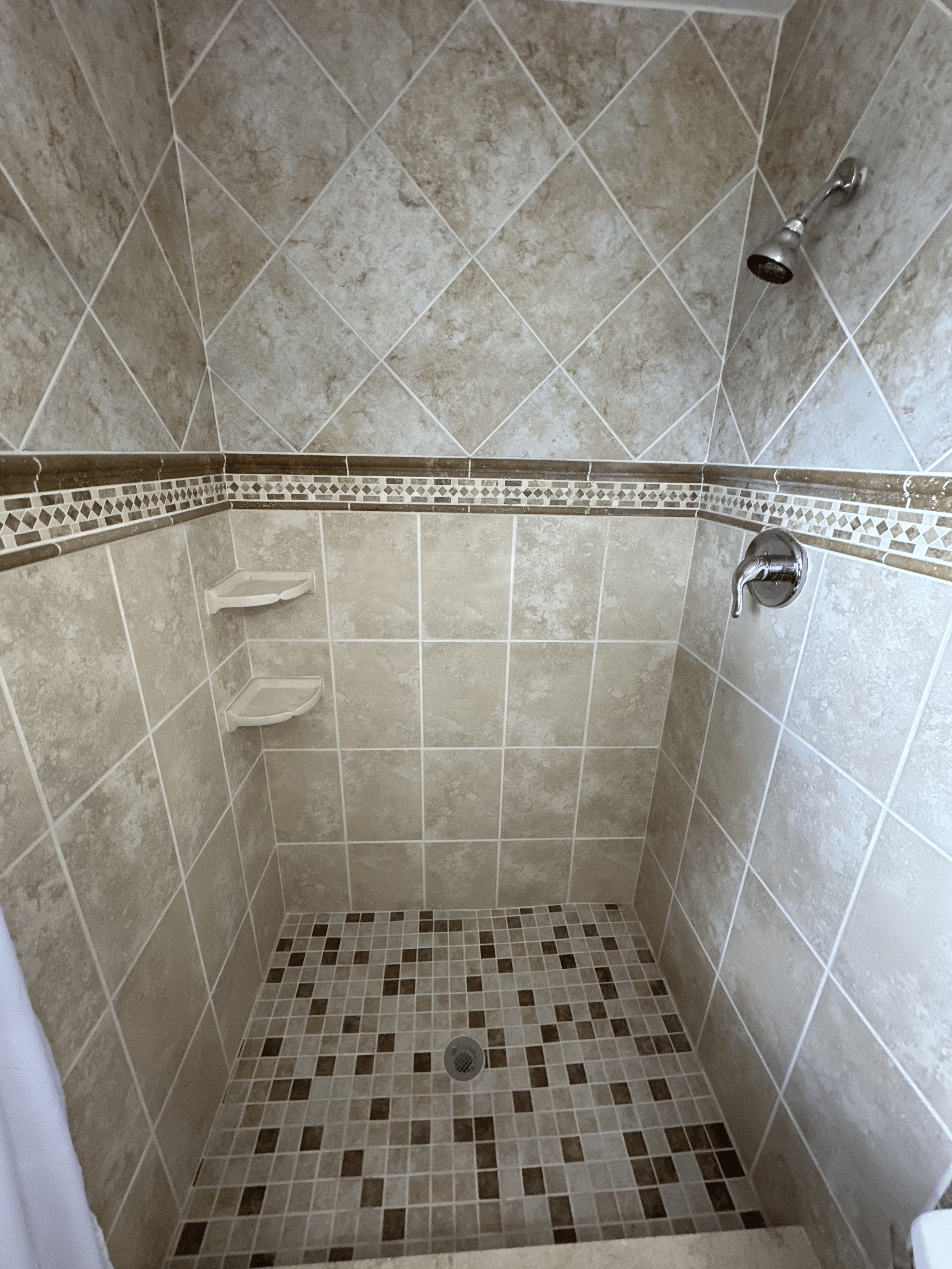 Shower stall with beige and brown tiled walls and floor, a wall-mounted showerhead on the right, two corner shelves on the left, and a drain in the center of the floor.