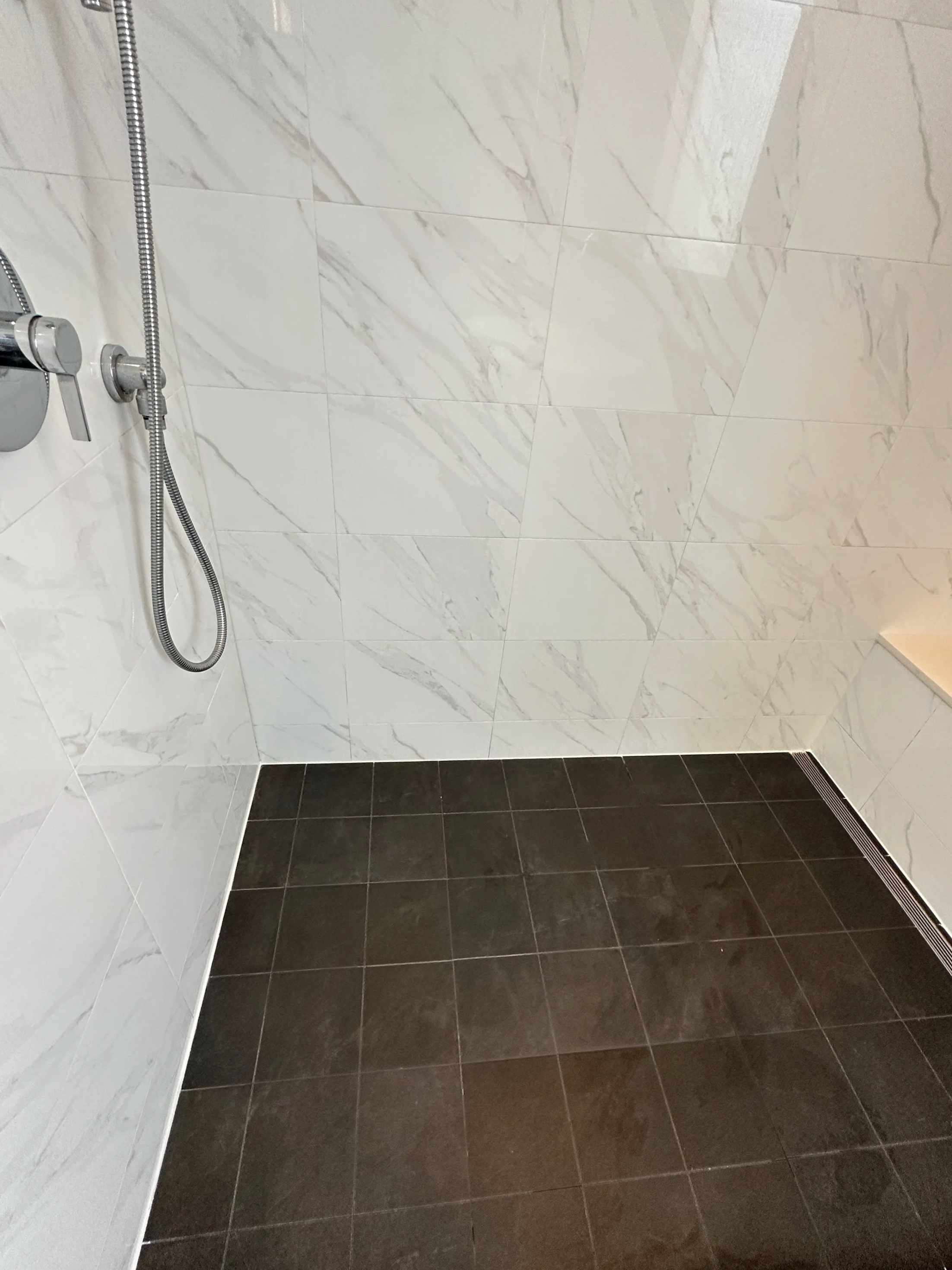 A bathroom shower area with white marble wall tiles, dark brown floor tiles, a silver showerhead connected to a flexible hose, and a floor drain.