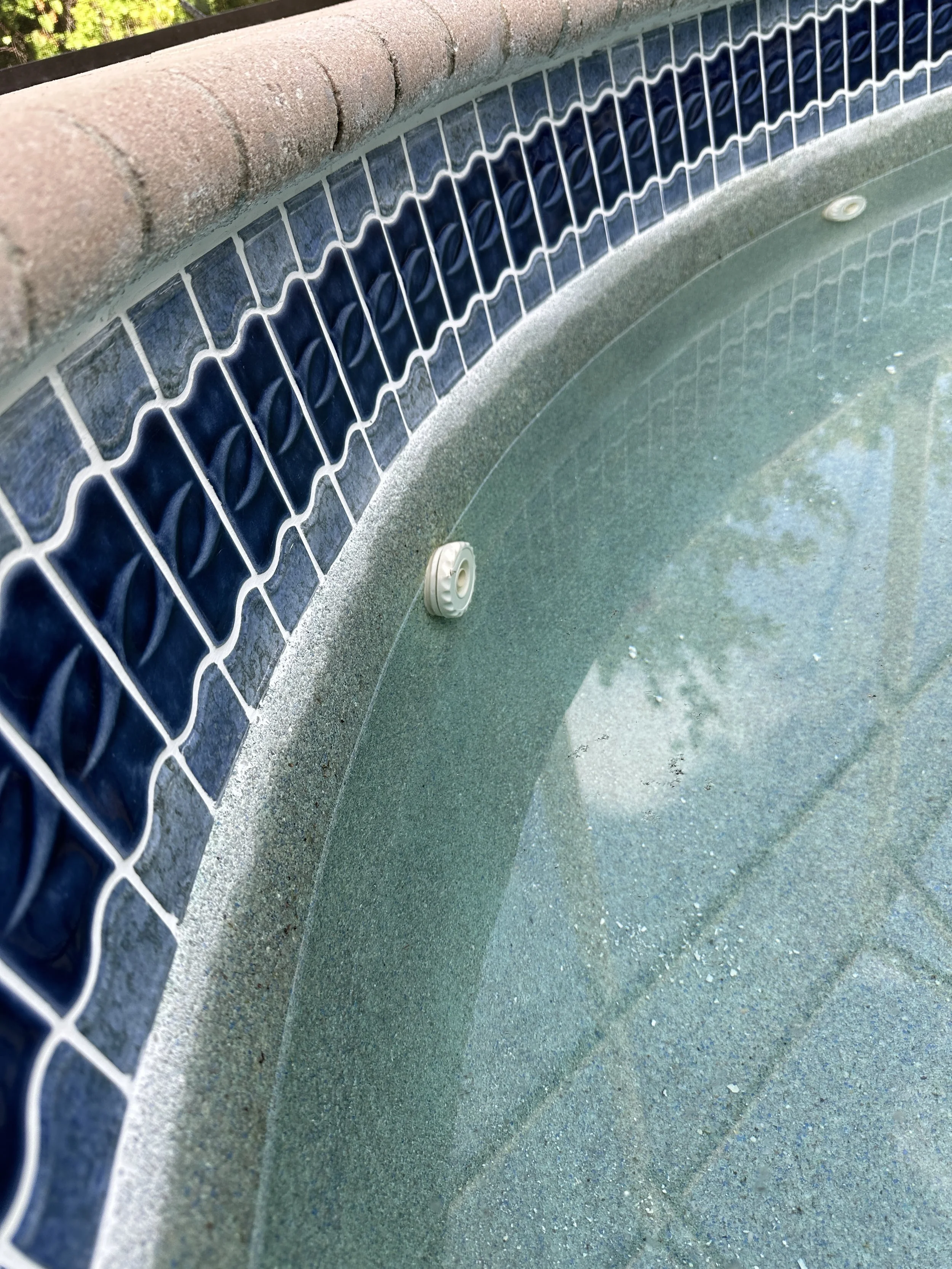 Close-up of the edge of a swimming pool with blue tiles, a brick border, and a metal drain or sensor in the pool.