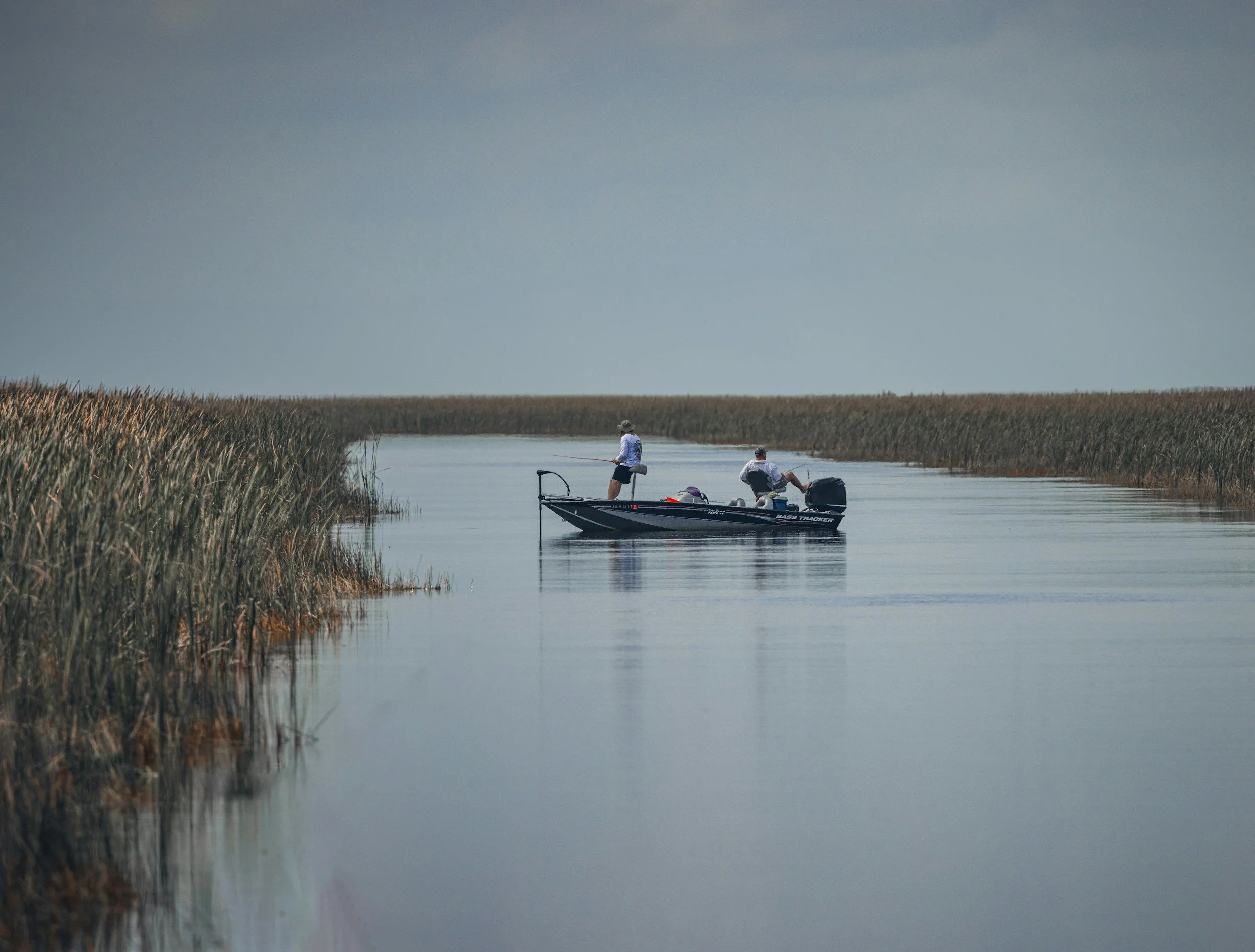 Two people fishing from a boat on a calm waterway surrounded by tall reeds and marsh plants under a slightly cloudy sky.