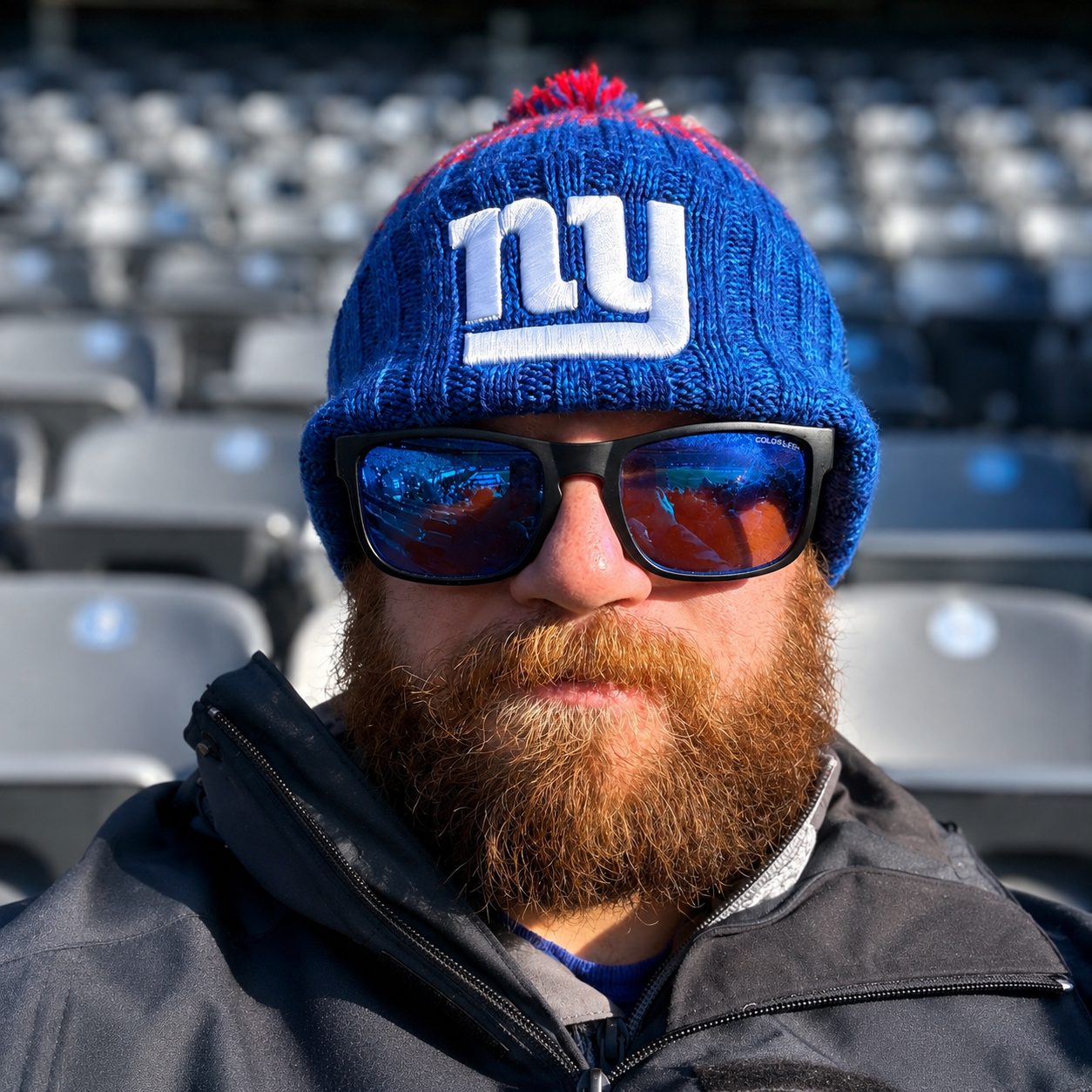Man with red beard wearing a blue NY Yankees knit beanie, sunglasses, and a black jacket, seated in a stadium with empty gray seats in the background.