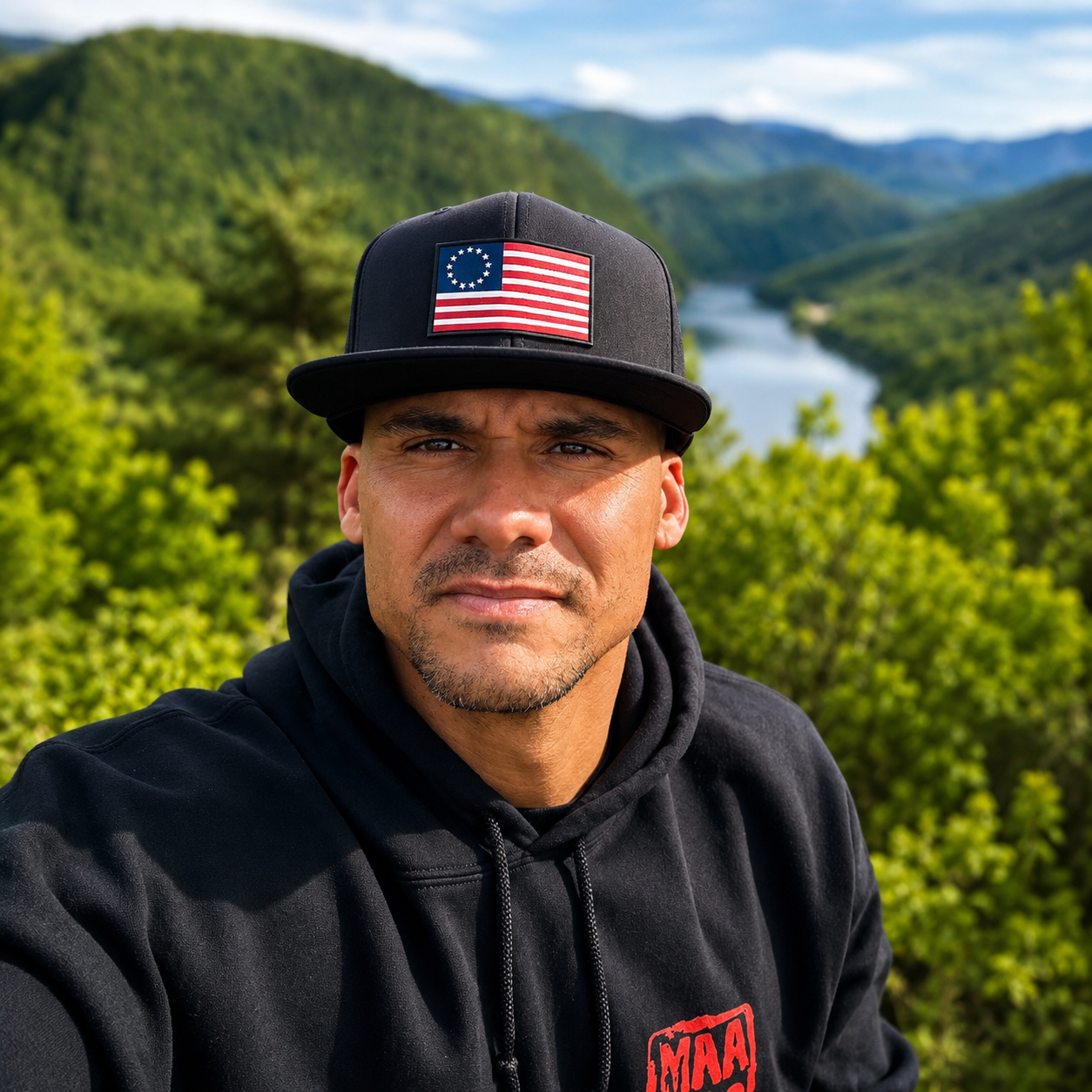 A man taking a selfie outdoors with a scenic background of mountains, a river, and green foliage. He is wearing a black hoodie and a black cap with an American flag patch.