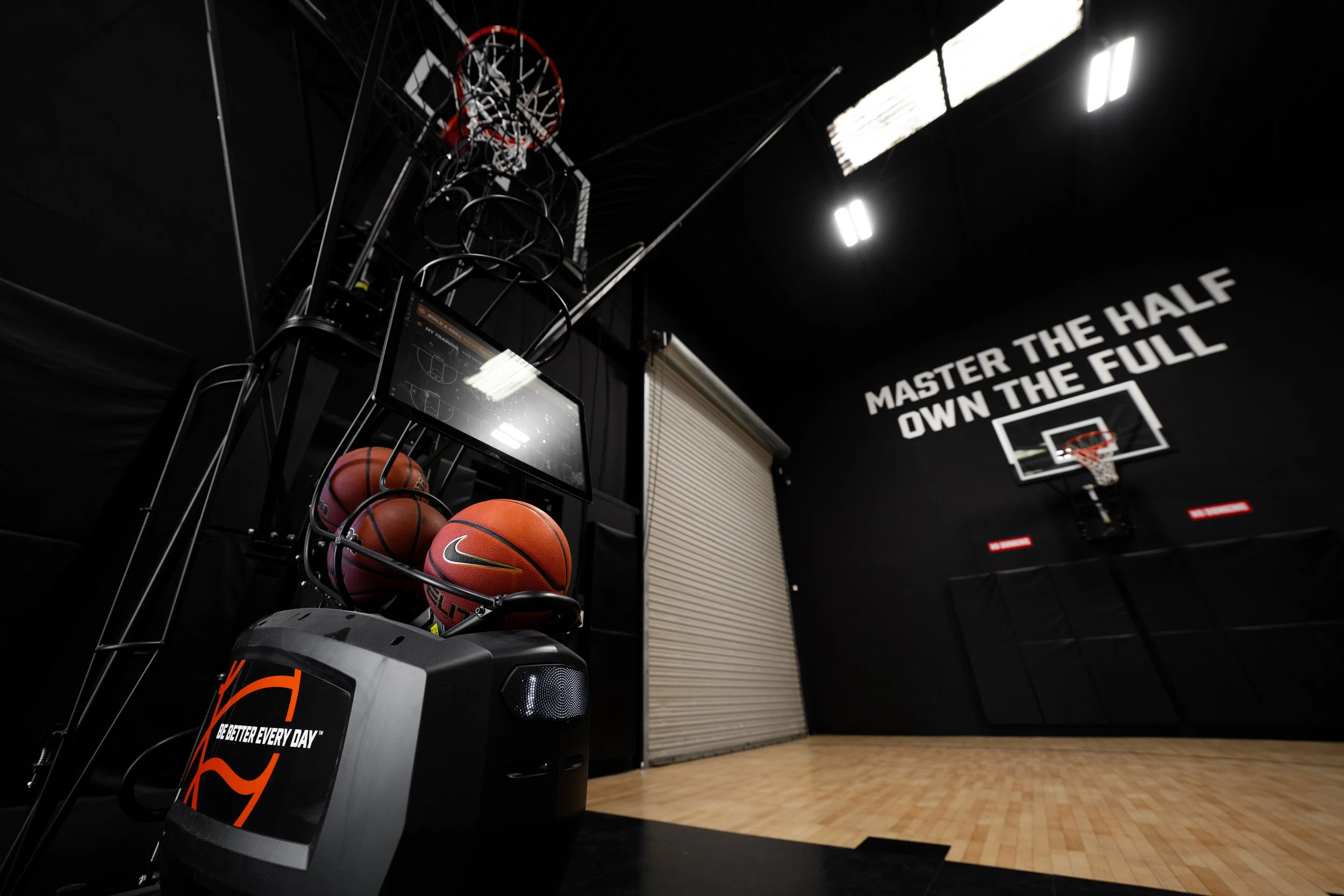 Basketball gym with a basketball shooting machine and basketballs, black walls with motivational quote, basketball hoop in the background, wooden floor.