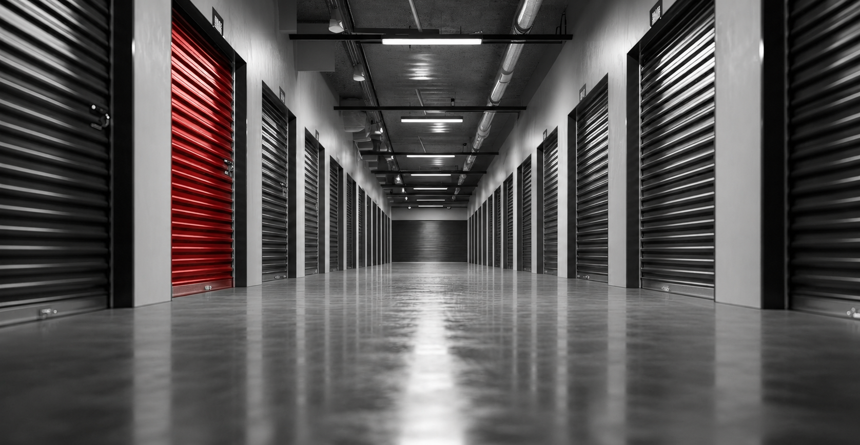 Long hallway in a storage facility. The image has a monochrome filter with a red accent. The ceiling has exposed pipes and modern lighting.