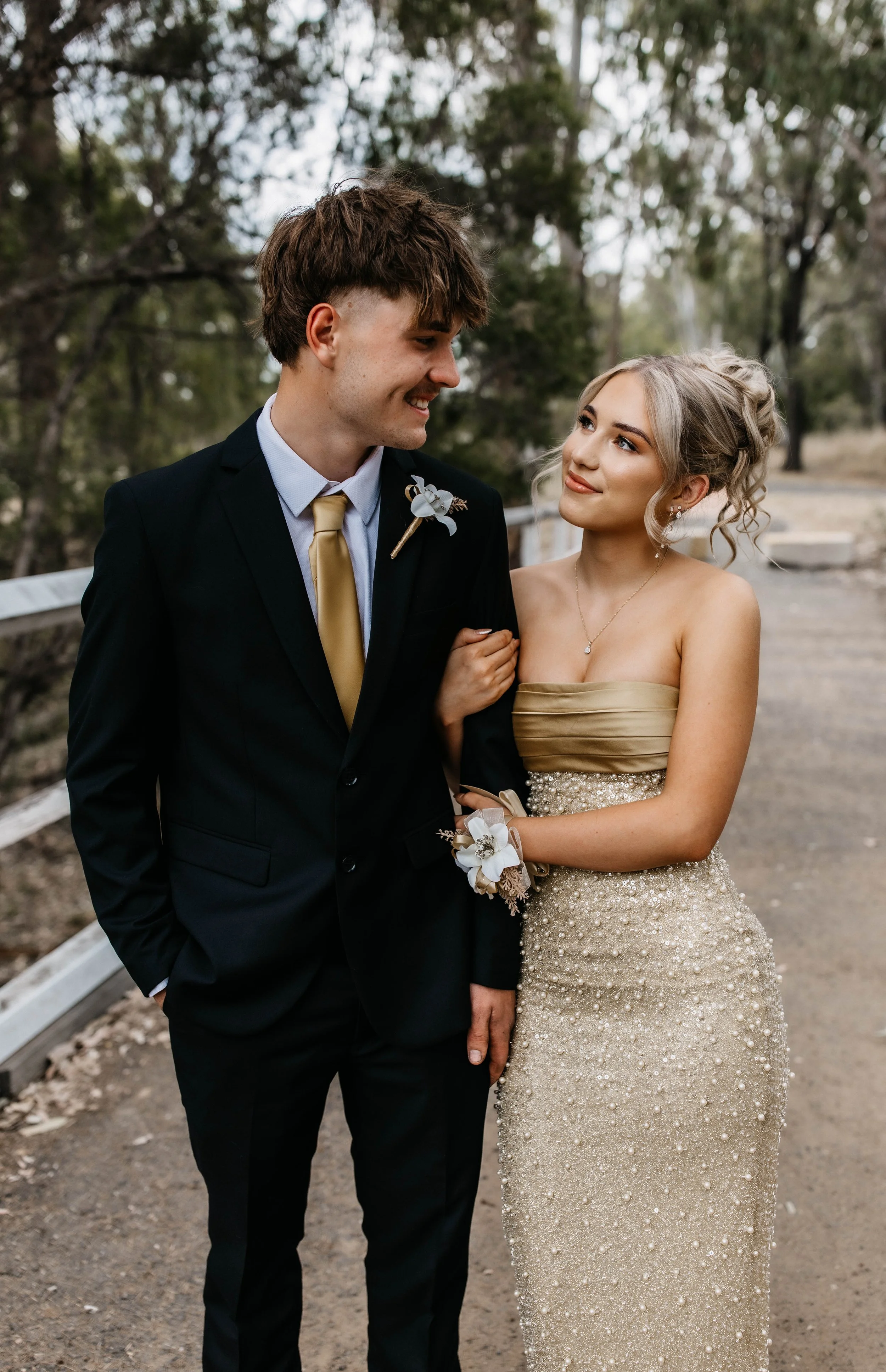 A young man in a black suit with a white shirt and gold tie, and a young woman in a strapless, beige, sparkling gown, standing arm in arm outdoors among trees. They are smiling and looking at each other.