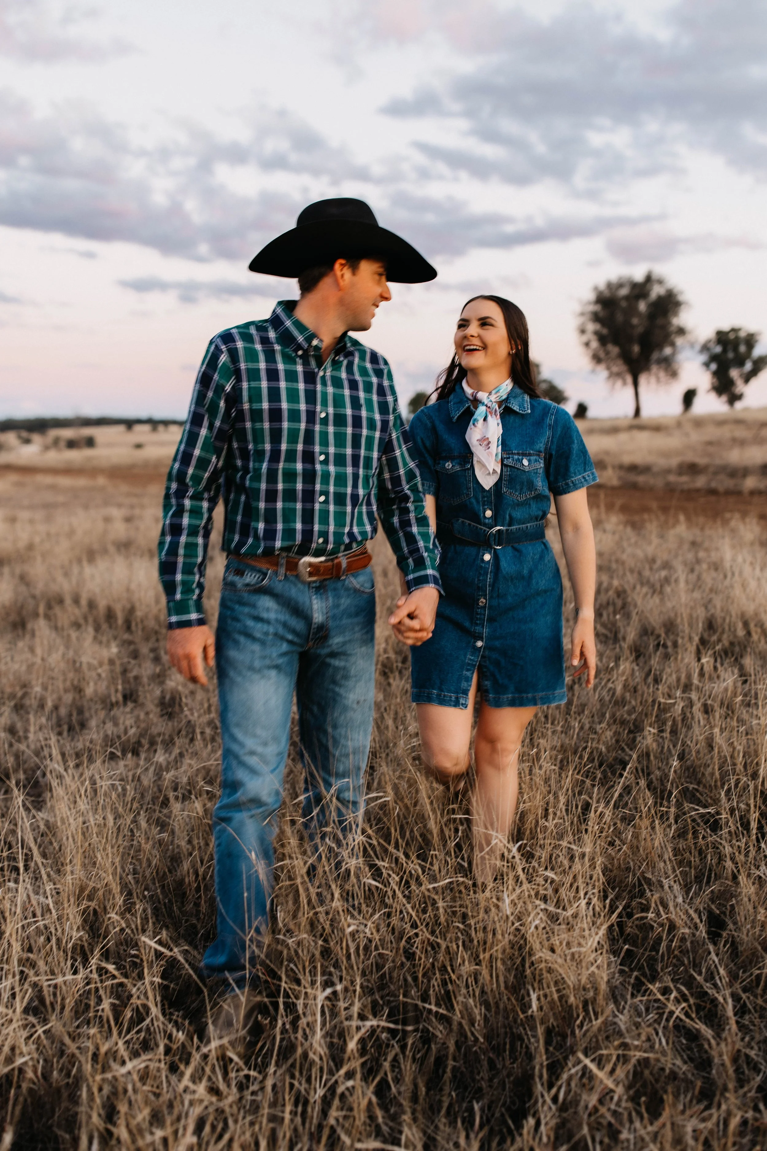 A young couple walking hand-in-hand through a field of tall, dry grass during sunset, smiling at each other.