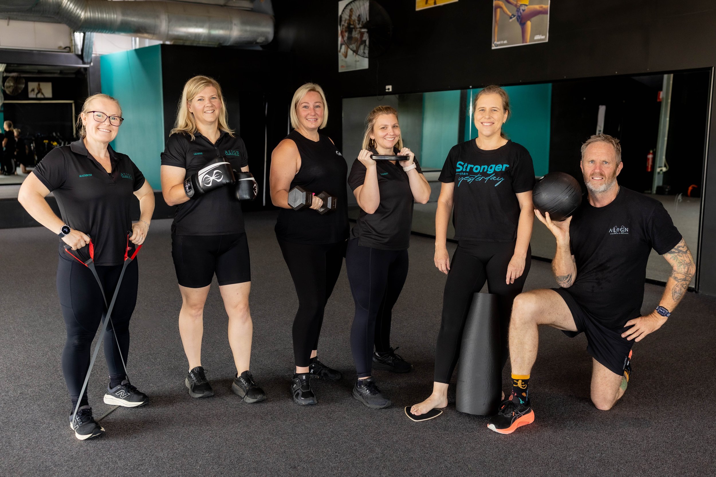 Group of six fitness instructors and workout enthusiasts in a gym, smiling and holding various exercise equipment, including dumbbells, a resistance band, a medicine ball, and a foam roller.