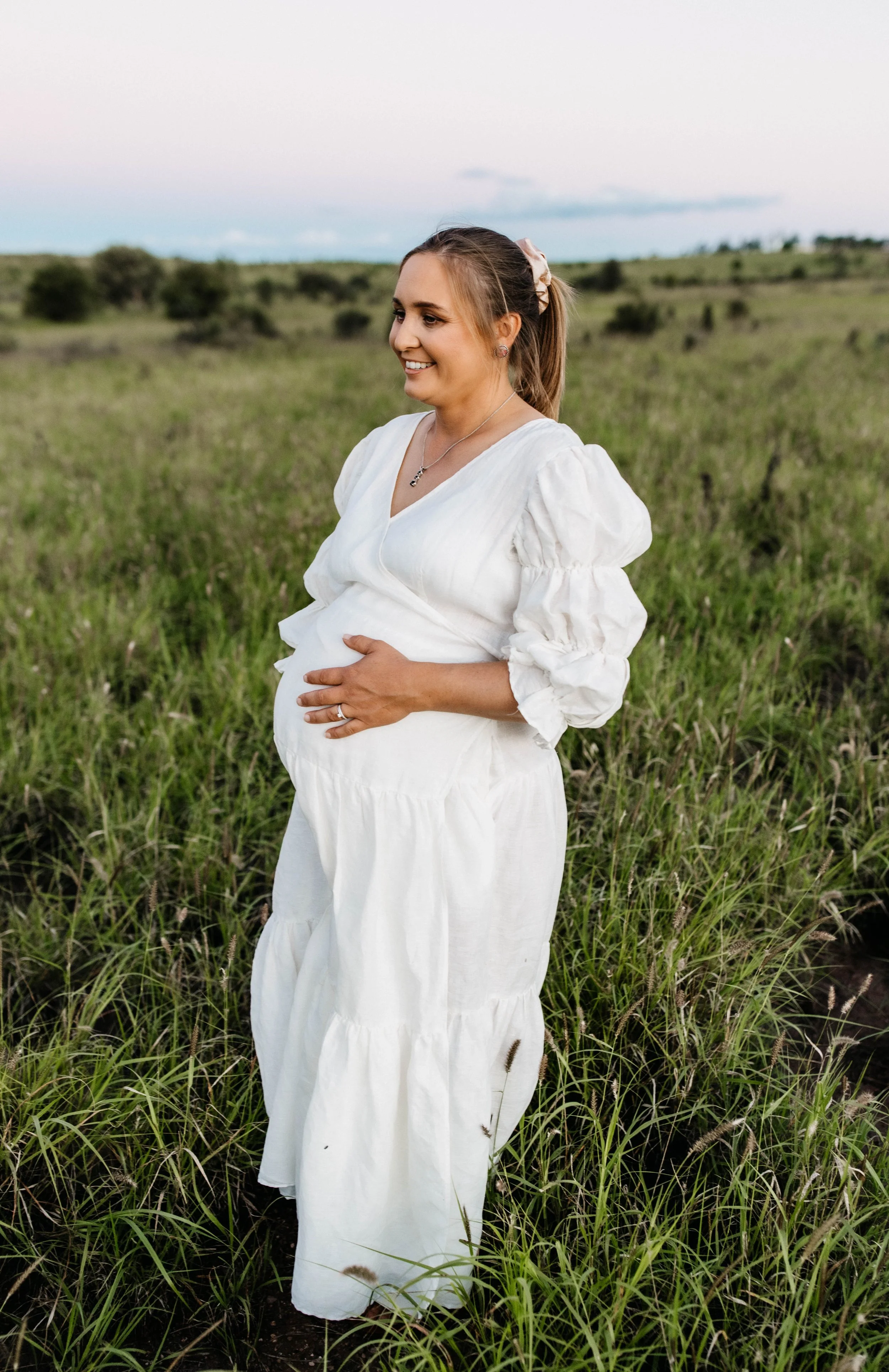 A pregnant woman dressed in a white dress standing in a grassy field, smiling and gently holding her belly with one hand, with a landscape of trees and a cloudy sky in the background.