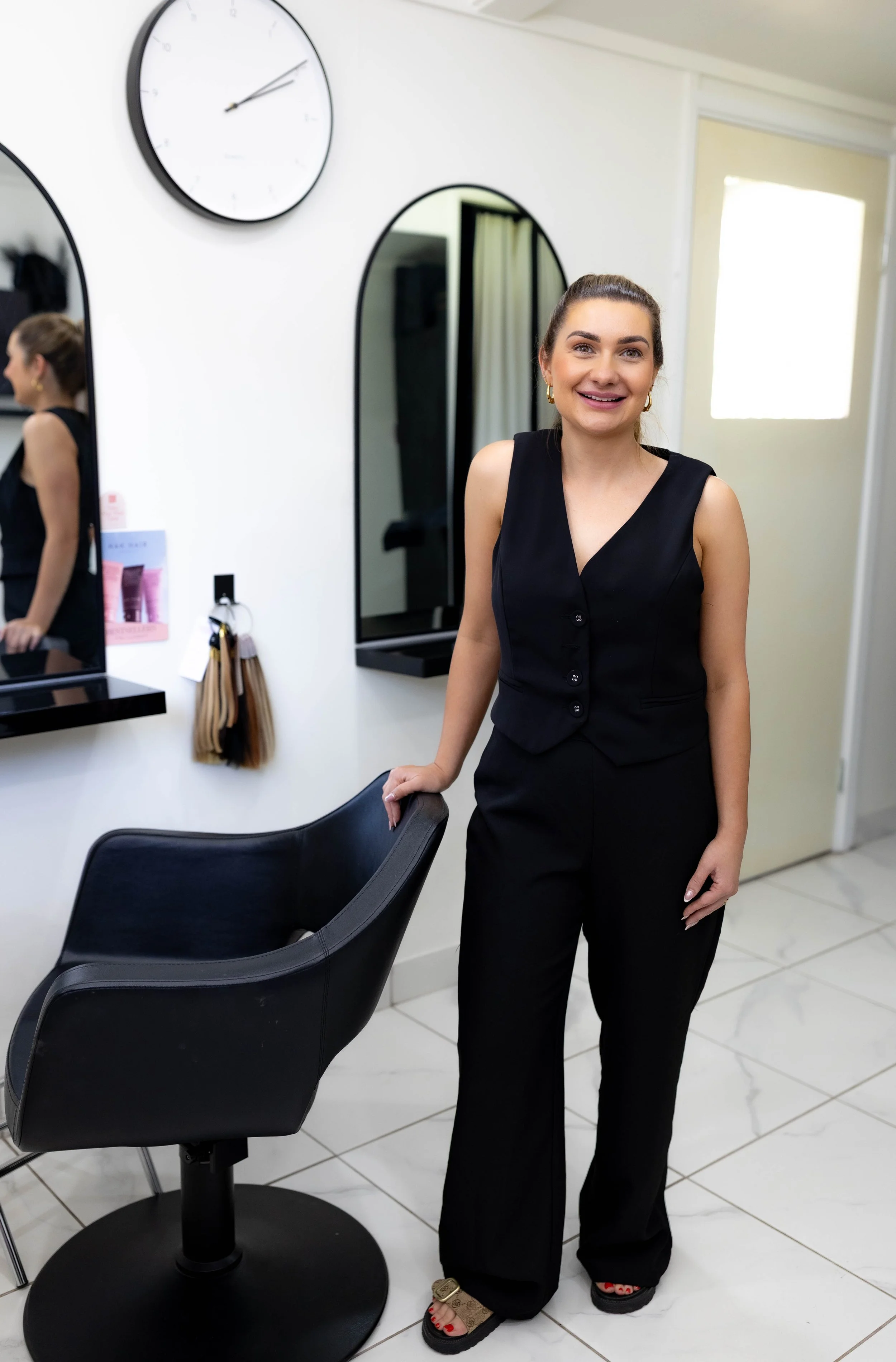 A young woman with dark hair in a ponytail, wearing a sleeveless black top, black pants, and sandals with a beige strap, standing in a salon or studio. She is smiling and looking at the camera. There is a black salon chair in front of her, and mirrors on the wall behind her, with a large clock showing 2:09.