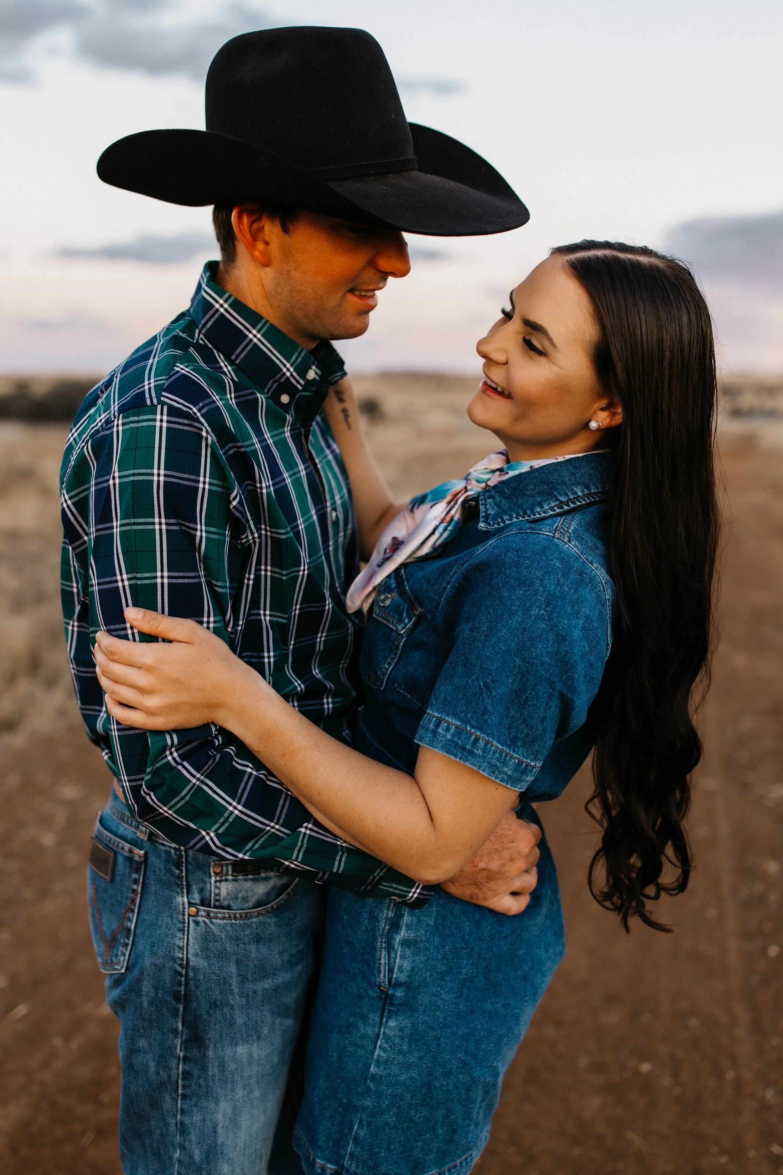 A couple embraces outdoors during sunset, smiling at each other with a cloudy sky in the background.
