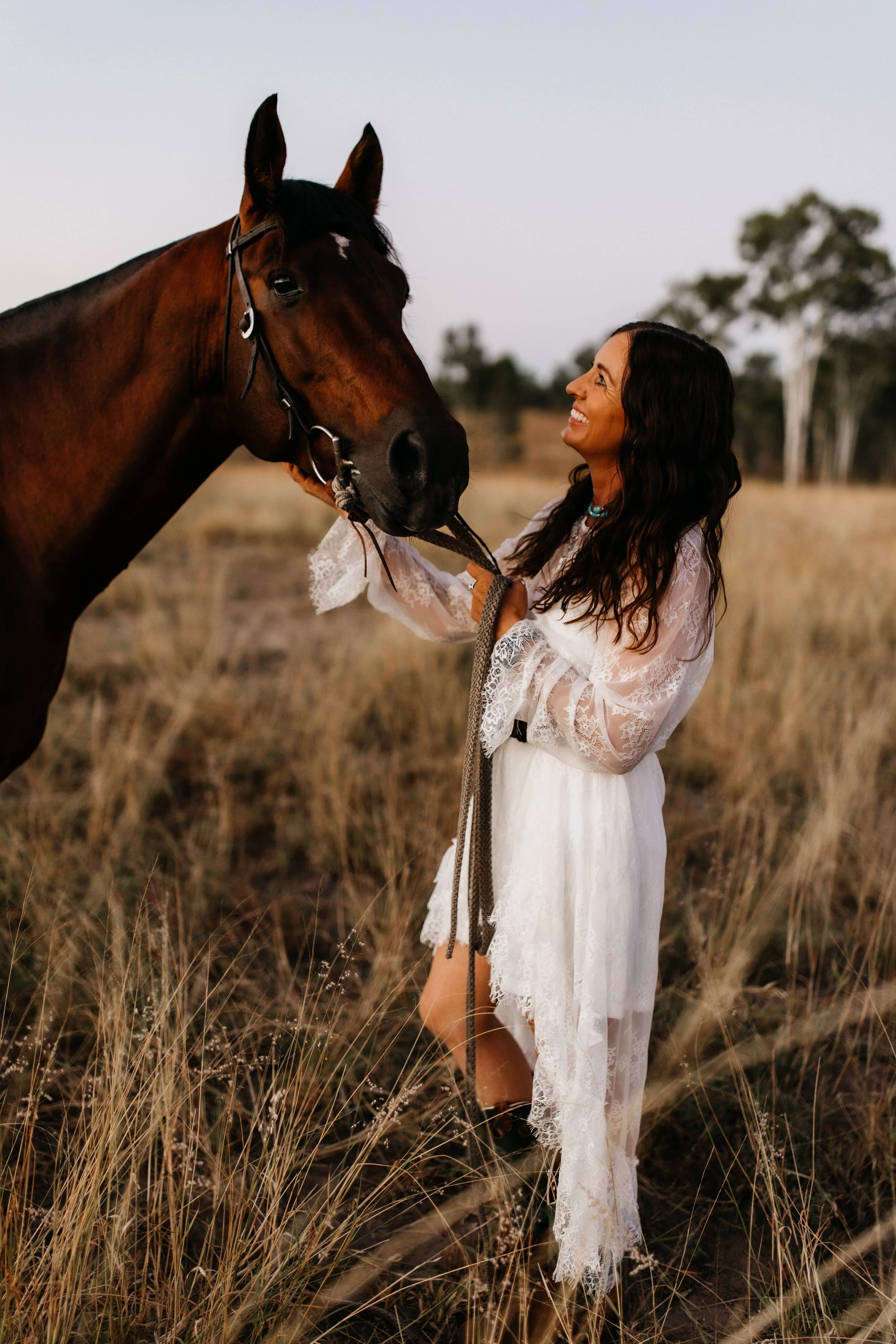 A woman with long dark wavy hair smiling at a large brown horse in a field of tall dry grass during late afternoon or early evening.