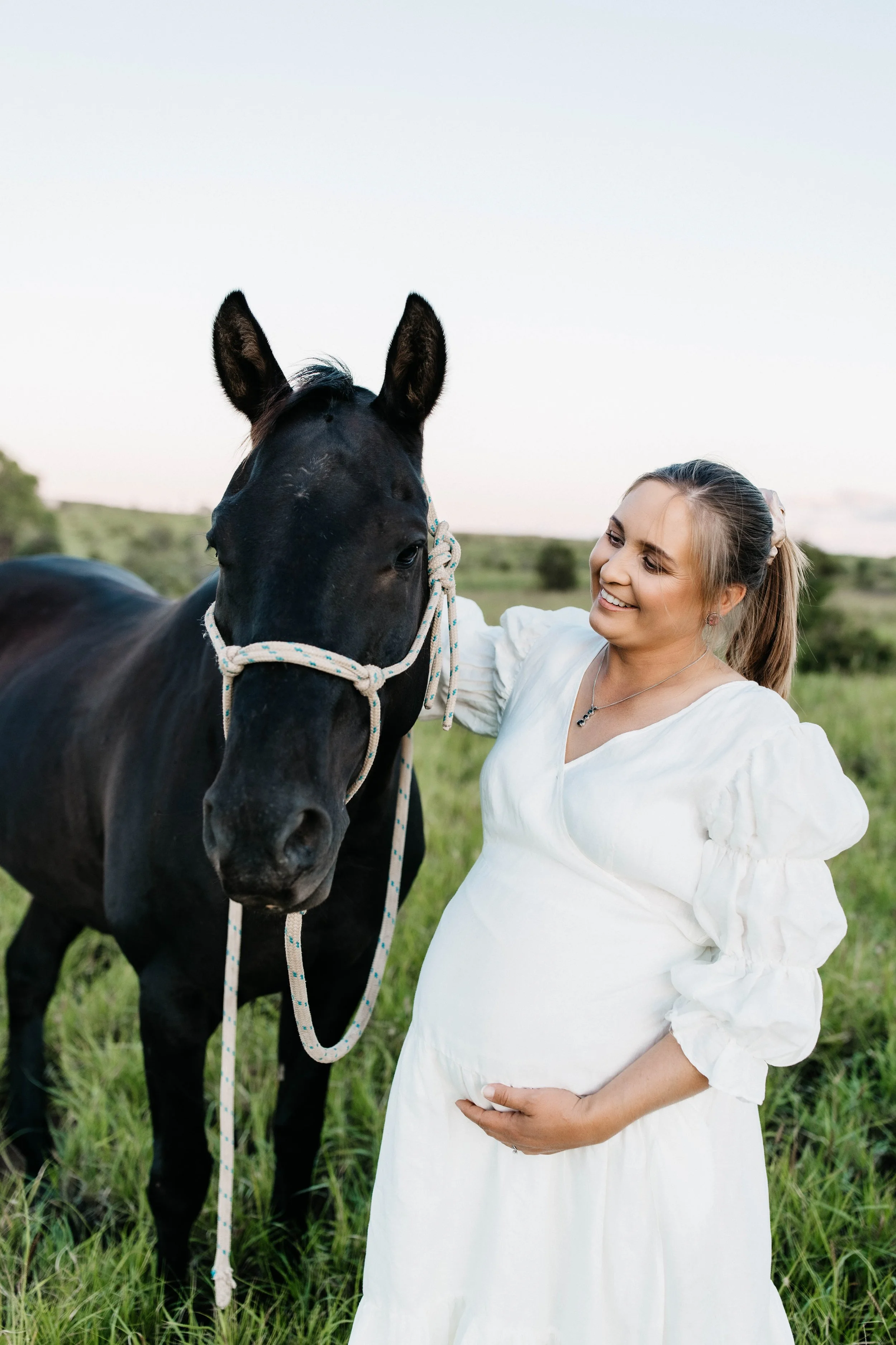A woman with blonde hair in a ponytail wearing a white dress standing in a grassy field, smiling at a black horse with a white rope halter.
